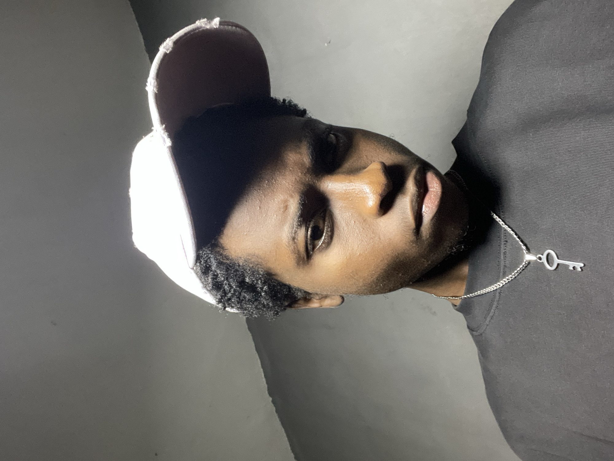 The media file shows a close-up, vertically oriented portrait of a young Black person wearing a light-colored baseball cap and a dark t-shirt. The subject is looking towards the camera with a neutral expression. A silver chain with a key-shaped pendant is visible around their neck. The background is a plain, textured wall with soft lighting, creating a shadow on the subject's face. The overall mood is calm and introspective. The image appears to have been taken indoors in Nkpor, Nigeria. There is no visible text in the image.