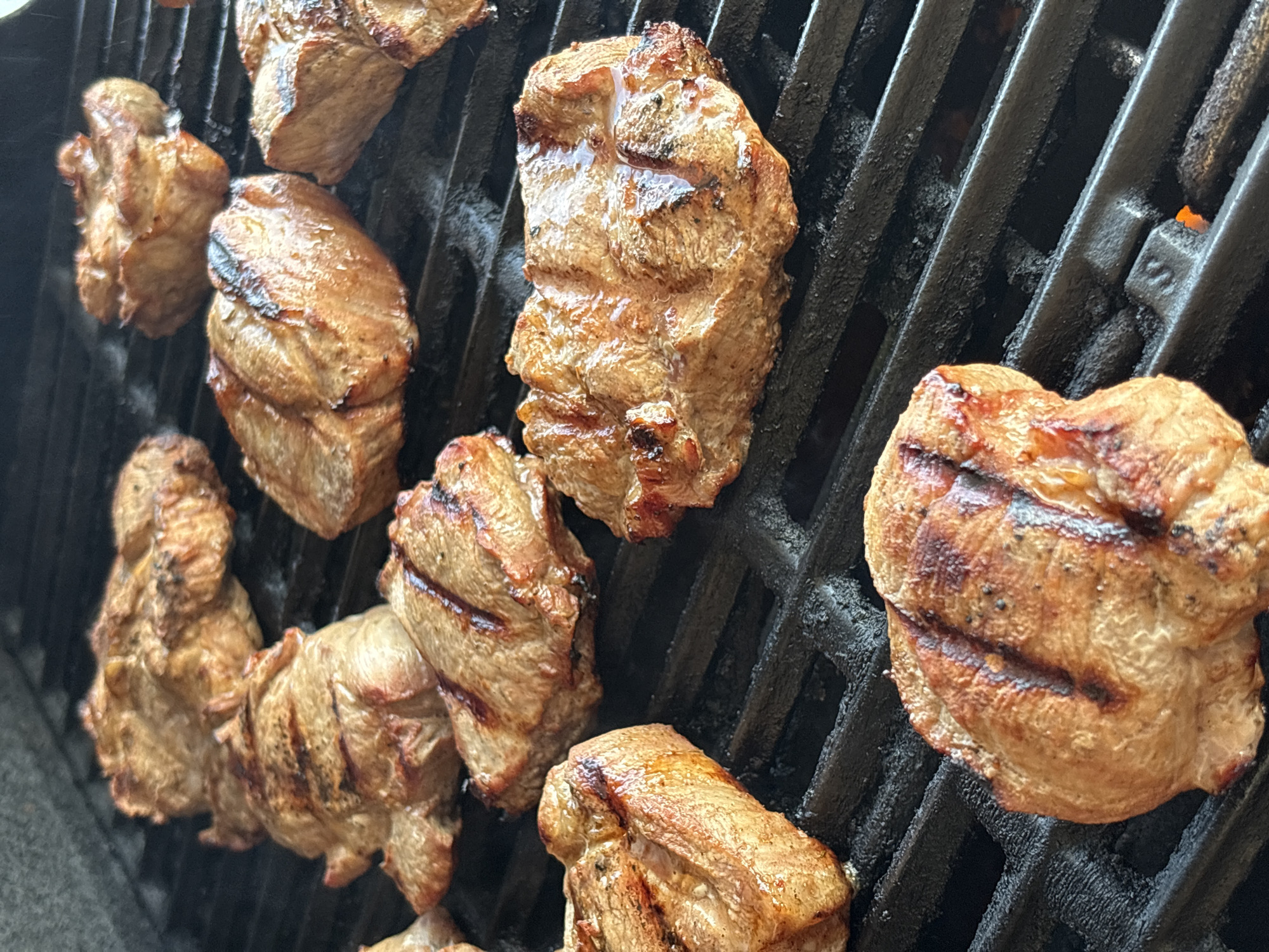 The image shows multiple pieces of grilled meat on a black cast-iron grill grate. The meat appears to be small, bite-sized portions, some stacked on top of each other. They have grill marks and a slightly browned, cooked exterior. Wisps of smoke are visible rising from the meat, indicating it is actively being cooked. The grill grate itself has some residual charring and grease. The background is out of focus, but appears to be dark, suggesting an outdoor setting, possibly a backyard or patio. The lighting is consistent with daylight. There is no visible text in the image. The scene depicts the process of grilling meat.