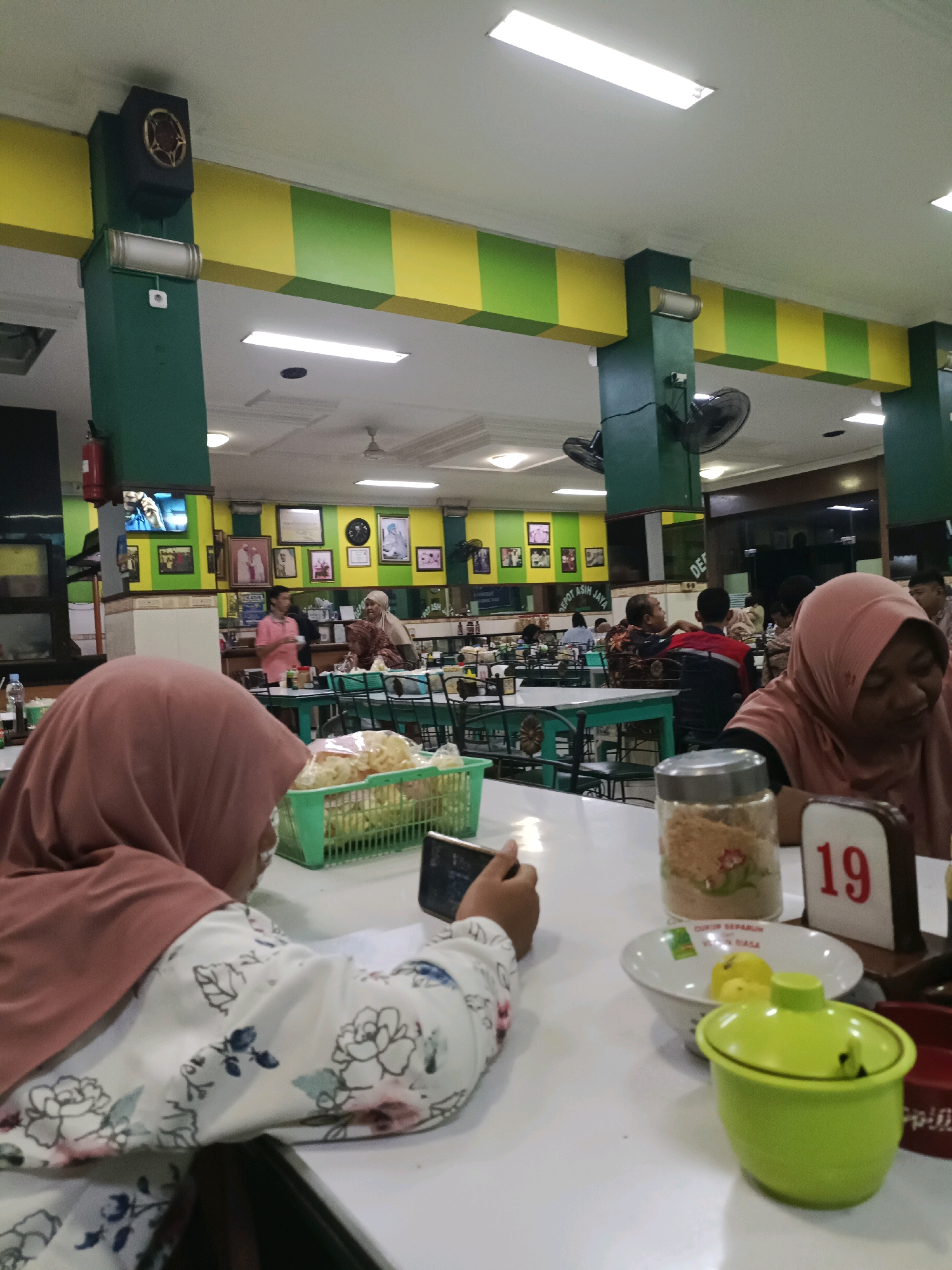 The image captures an indoor dining area, likely a restaurant or food stall in Dapur, Indonesia. The decor features green and yellow checkered beams supporting the ceiling, with fluorescent lights illuminating the space. Several individuals, predominantly women wearing hijabs, are seated at tables. In the foreground, a woman in a patterned outfit is engrossed in her mobile phone. On the table, there's a green basket with snacks, a bowl with yellow food items, a jar of condiments, and a table number display showing "19." In the background, other patrons are visible, some interacting while others appear to be eating. The environment suggests a casual dining setting with multiple tables and chairs. The walls are adorned with framed pictures and a television screen displaying an image. Fans are mounted on the ceiling and columns to circulate air. A fire extinguisher is mounted on a green column.