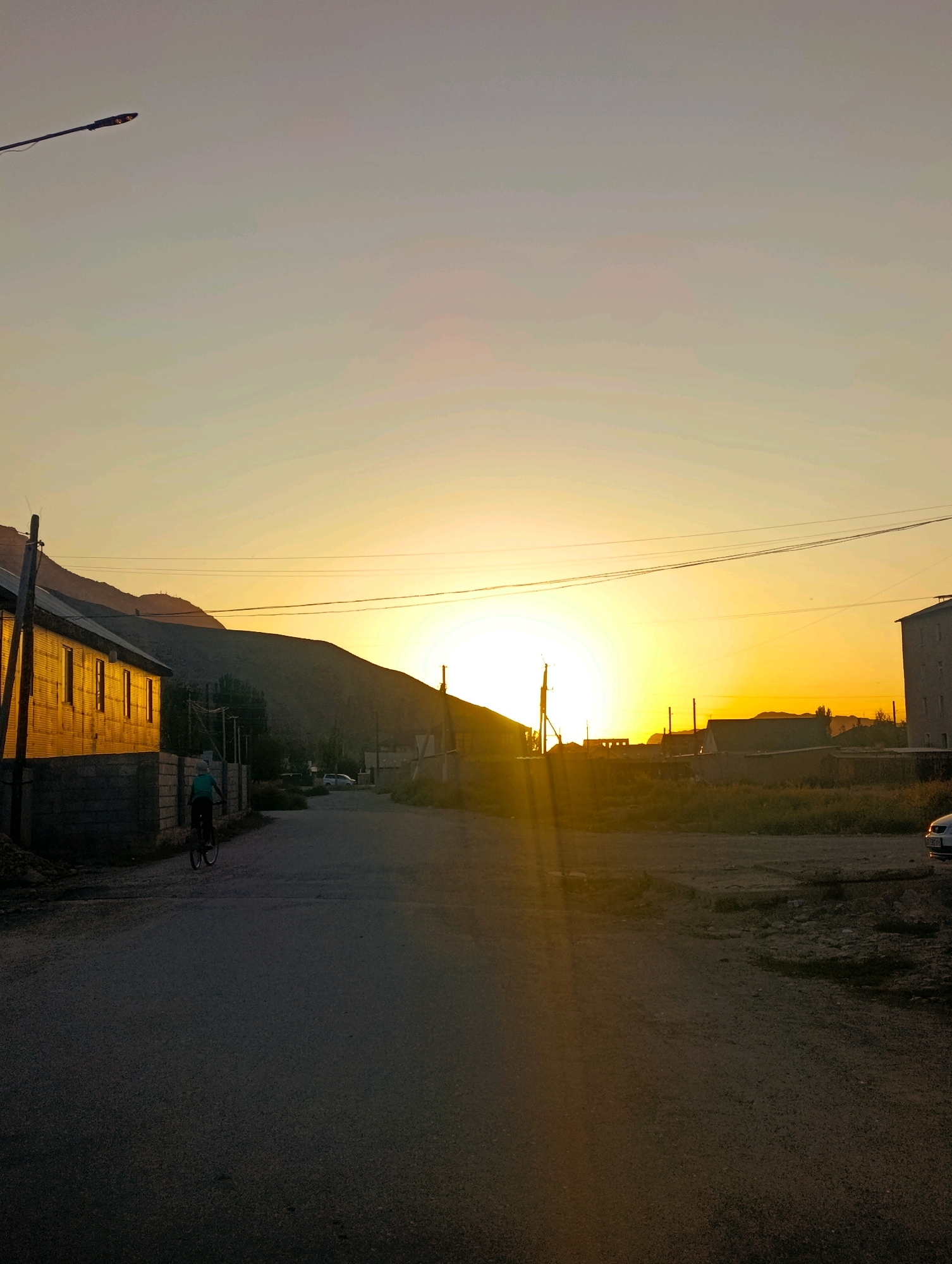 The image depicts a rural street scene in Naryn City, Kyrgyzstan, during sunset. A person is riding a bicycle down a paved road that stretches into the distance. The road is lined with low-rise buildings on the left, made of brick and corrugated metal, and undeveloped land with some vegetation on the right. In the background, hills rise up, silhouetted against the vibrant orange and yellow hues of the setting sun. Power lines crisscross the sky, and a street lamp is visible on the left. The overall atmosphere is serene, with the golden light of the sunset casting long shadows.