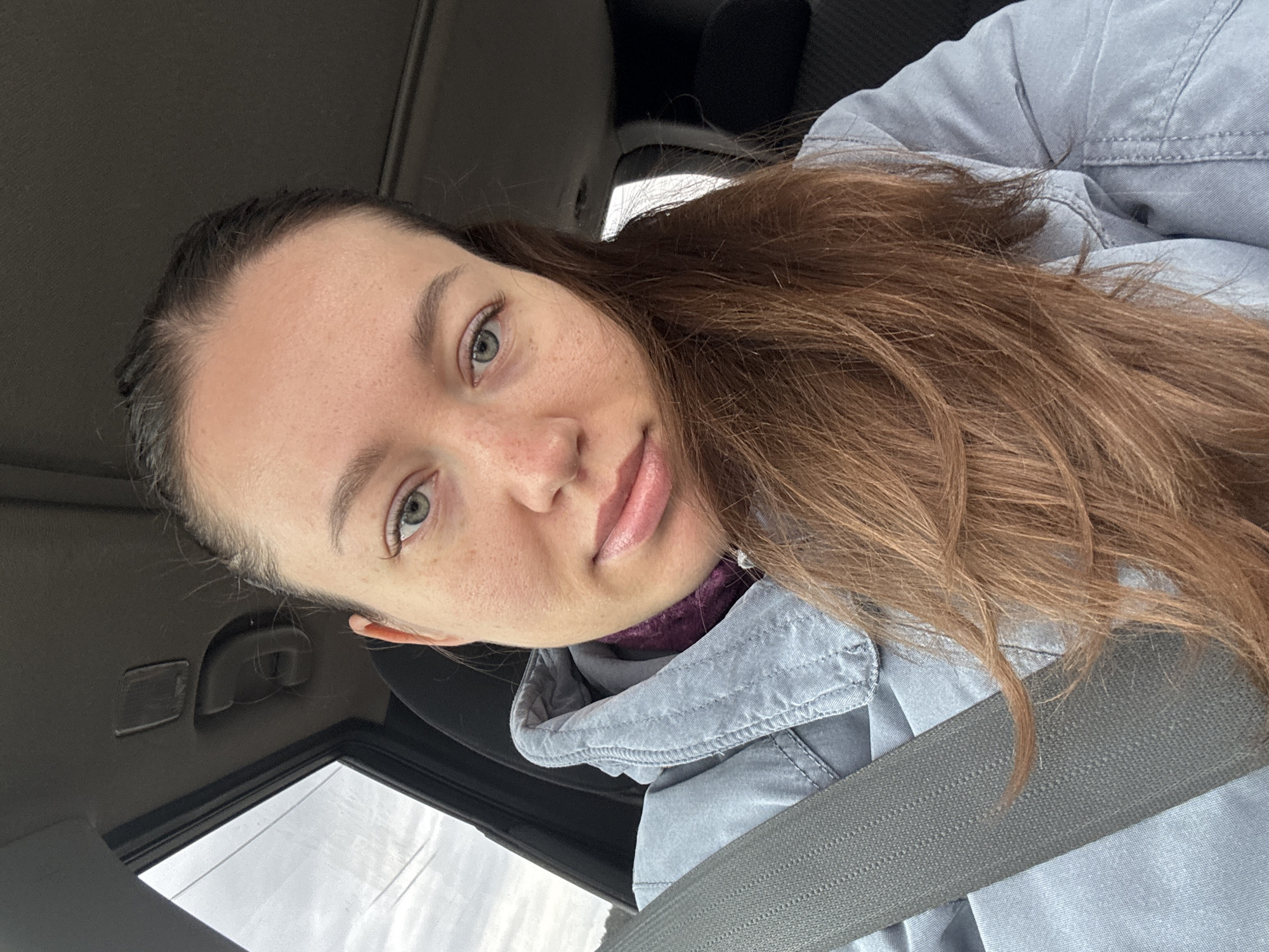 A woman is seated in a vehicle, wearing a light blue jacket and a seatbelt. She has brown hair, fair skin, and light-colored eyes, and is looking at the camera. The interior of the car is visible, including the roof and a window. The location in the city of Lipaki, Russia is undetermined.