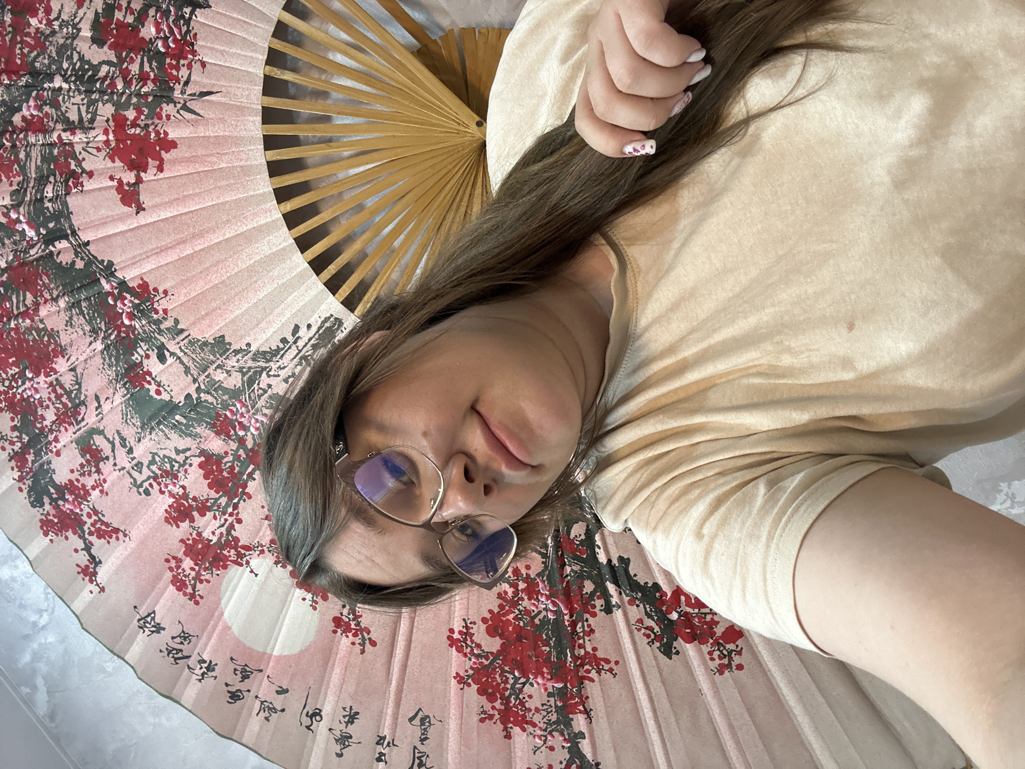 A woman lies on a patterned fan, positioned on a light gray surface. The fan is two-toned, combining a light pink section adorned with red cherry blossoms and black calligraphy with a natural-colored wood section composed of evenly spaced slats. The woman wears gold-rimmed glasses and a tan-colored t-shirt. Her right hand is visible near the top right corner of the image. The photo appears to be a selfie taken from a high angle. It is not possible to determine the location based on the image.