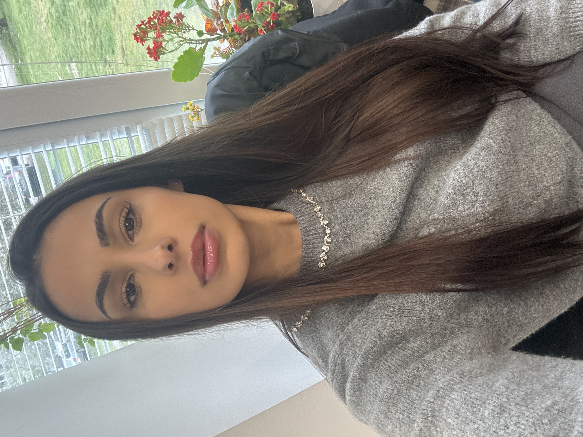 A young woman with long, dark brown hair is looking directly at the camera. She is wearing a gray sweater and a delicate silver necklace. The background includes a window with white blinds and a view of greenery, including plants with red and yellow flowers. A black jacket is draped behind her. The image does not provide enough information to determine the specific location in Sofia, Bulgaria.