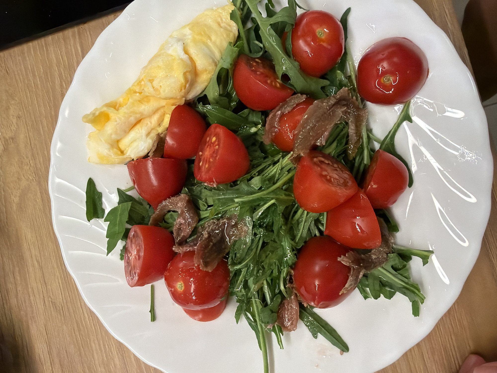 A white, irregularly shaped plate holds a meal, centered on a light-colored wooden table. The dish primarily features a salad base of fresh, dark green arugula leaves, interspersed with bright red cherry tomatoes. Some tomatoes are whole, while others are halved, revealing their internal structure. Small, brownish-silver fillets, consistent with anchovies, are scattered on top of the arugula and tomatoes. To the upper left side of the plate, a portion of pale yellow, folded or rolled eggs, resembling a small omelet or scrambled eggs, completes the meal. The plate itself has subtle, raised decorative lines on its rim, especially visible towards the bottom right. The background is mostly the wooden surface, with a dark, out-of-focus element appearing in the upper left corner. No people or text are visible, and the time of day is not discernible. The meal is presented, ready for consumption in what is noted to be Budva, Montenegro.