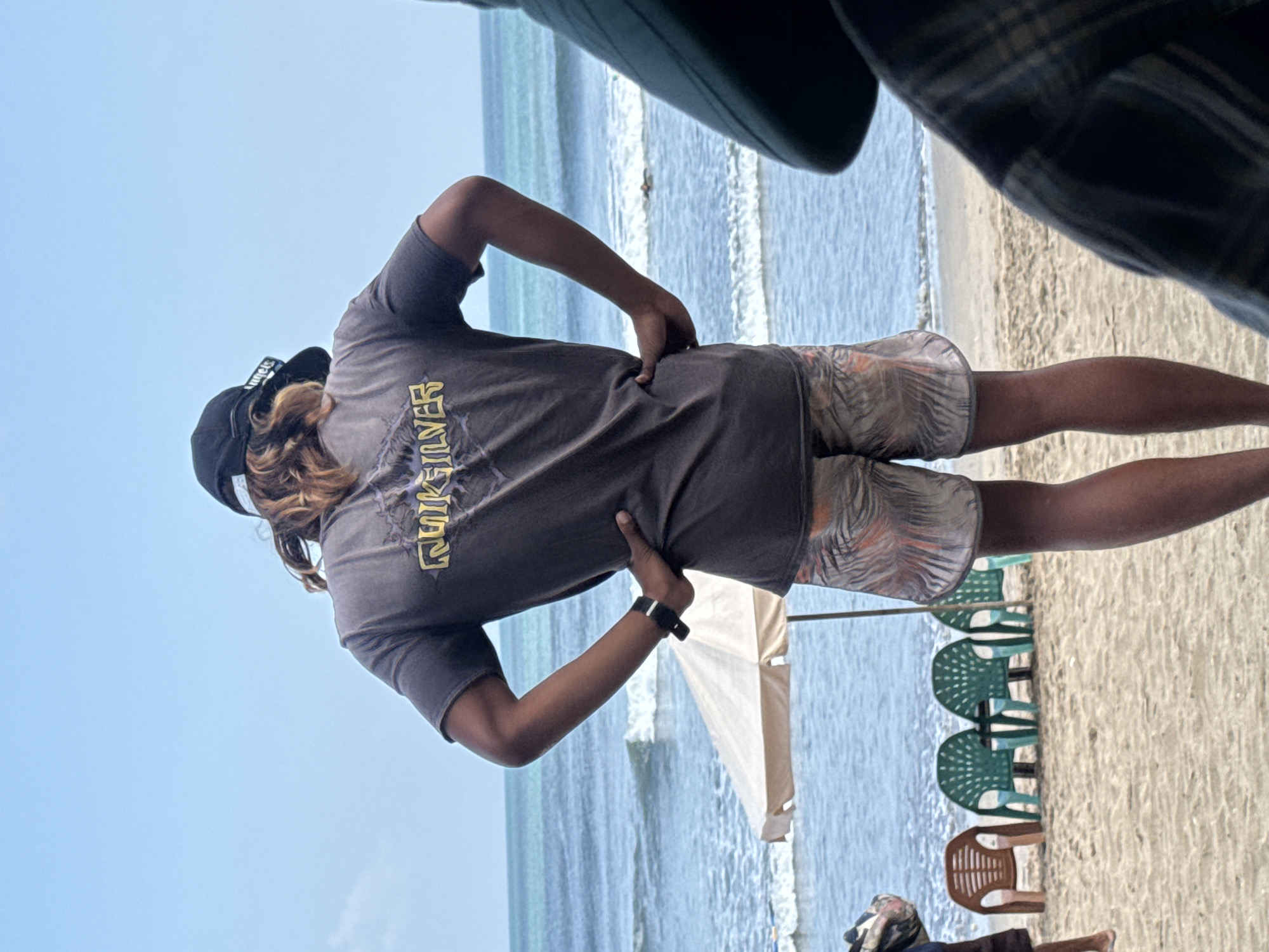 A medium shot captures a person standing on a sandy beach, facing away from the camera towards the ocean. The person is wearing a dark t-shirt with a "Quiksilver" logo in yellow text across the back and patterned shorts. They have their hands on their hips. Behind them, the ocean has moderate waves under a clear blue sky. To the left, a light-colored beach umbrella is partially visible, and several plastic chairs are arranged on the sand. In the distance, a small figure can be seen in the water. The scene appears to be in Kuta, Indonesia.