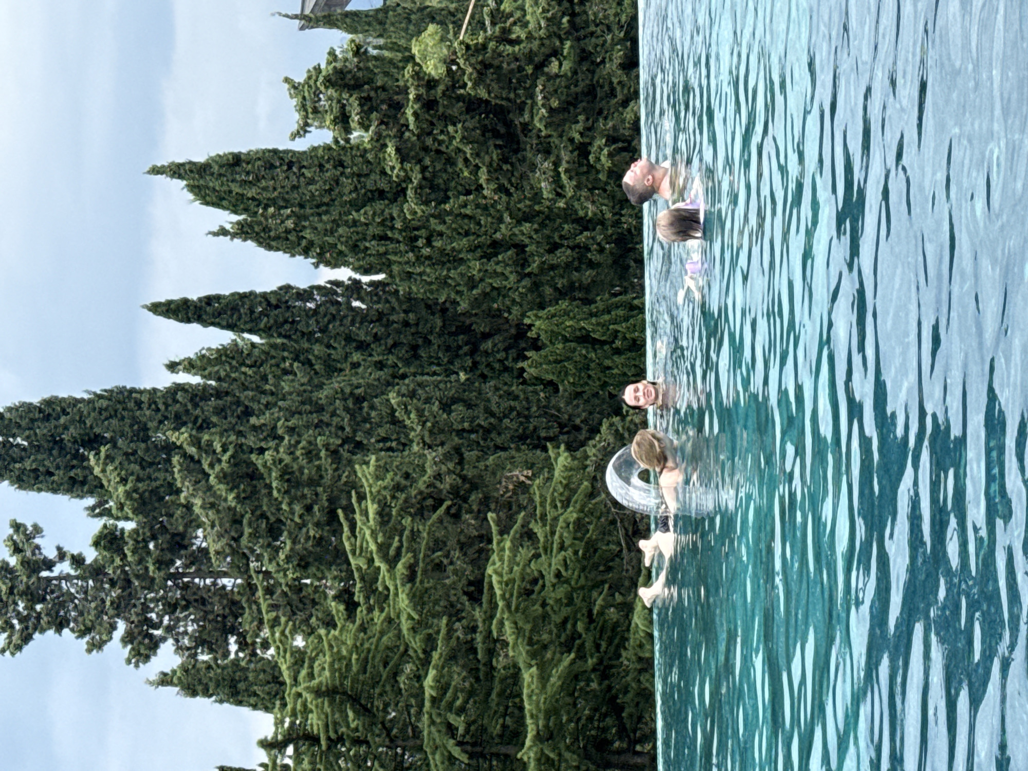 The image shows a group of people swimming in an infinity pool. The pool is surrounded by lush green trees, and the sky is a pale blue. There are at least three people visible in the water. One person is a man with short hair, looking up towards the sky. Another person appears to be a child with blonde hair, wearing a purple swimsuit and holding onto a clear inflatable ring. A third person is partially visible at the bottom of the frame, their face also turned upwards. The water is clear and reflects the light, creating a shimmering effect. The trees are tall and dense, suggesting a natural or resort-like setting. The image appears to be taken during the daytime, with good lighting. The overall mood seems relaxed and joyful, as the individuals are enjoying themselves in the water. The location is stated to be Yalta, Ukraine.