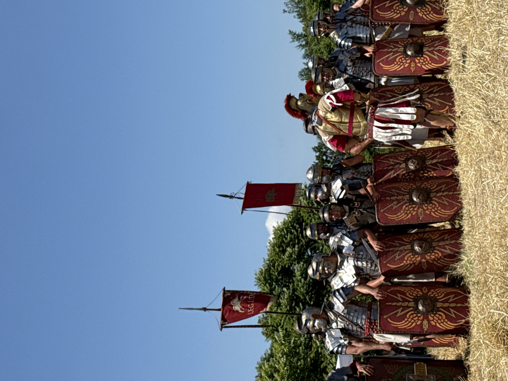 A group of approximately twenty individuals, dressed as Roman legionaries for a historical reenactment, stands outdoors in a sunny field of dry, golden straw in Gigen, Bulgaria. The figures wear metallic segmented armor (lorica segmentata), helmets, and red and white tunics. Each prominently displays a large, rectangular scutum (shield) adorned with intricate gold winged lightning bolt patterns surrounding a central metallic boss in the shape of a lion's head. A central figure in the foreground wears elaborate red and gold armor with a distinctive red-plumed helmet. In the background, a line of green trees is visible beneath a clear blue sky, with two military standards positioned above the foliage. The left standard is a red vexillum featuring a white horse or Pegasus and the partial text "LEG I ITA". The right standard, also red, displays an indistinct animal emblem.