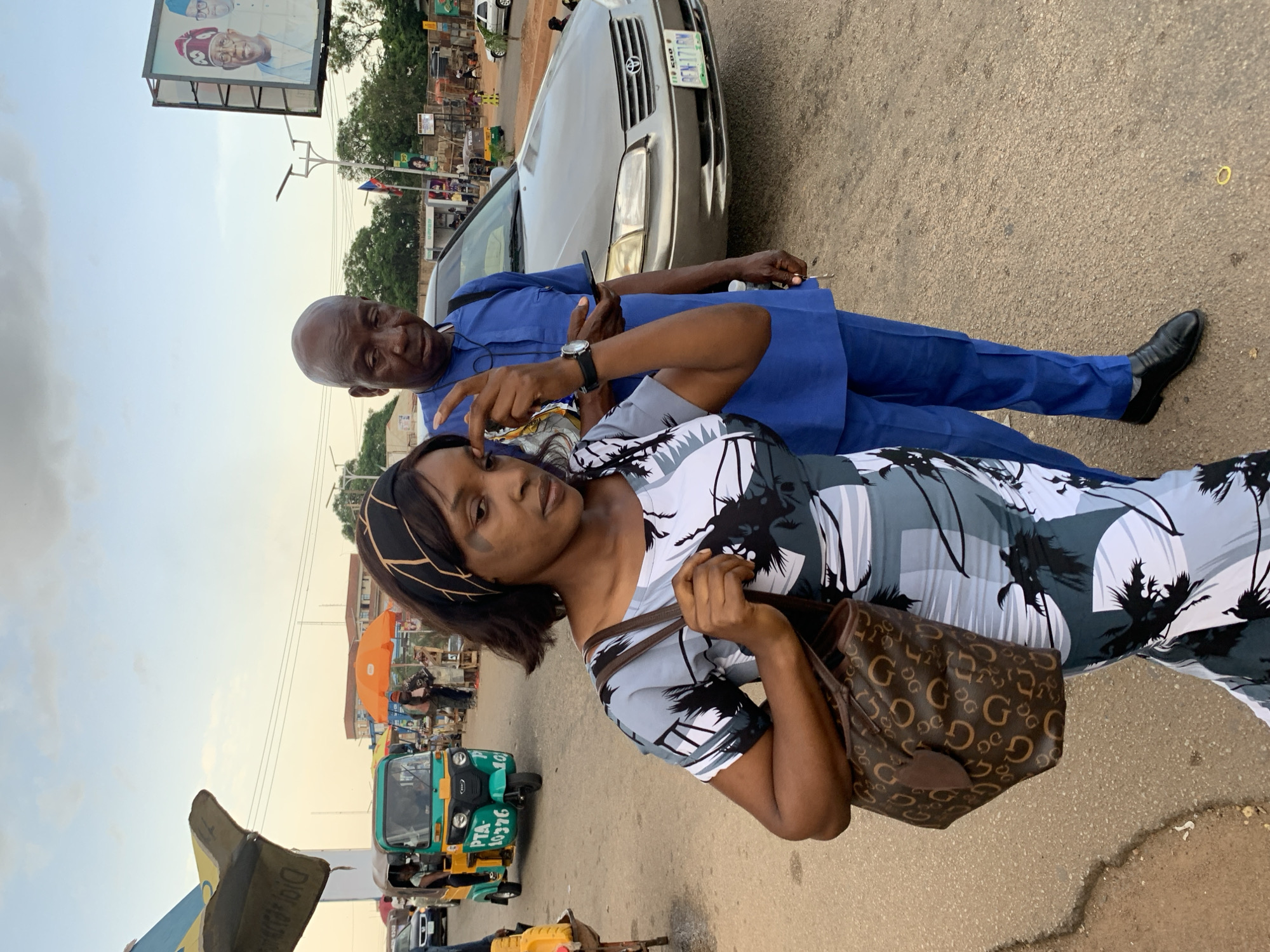 In an outdoor, urban setting, potentially in Jos, Nigeria, a woman and a man stand on a paved road. The woman, in the foreground and to the left, wears a patterned dress with black, white, and grey palm tree motifs and a black headband. She carries a brown designer handbag with a repeating "GG" logo and a small brown tag. Her right hand is raised towards her face, and she appears to be touching her eyebrow. The man, to the woman's right and slightly behind her, is wearing a blue traditional Nigerian outfit consisting of a long-sleeved shirt and trousers. He holds a smartphone in his left hand and is looking down. In the background, a silver sedan car is parked on the road. Further in the background, there are market stalls with orange umbrellas, a three-wheeled auto-rickshaw (Keke Napep), and a large billboard featuring portraits of two men in traditional attire. The sky is visible with some clouds, suggesting it is late afternoon or early evening. The overall scene depicts a moment on a busy street with elements of commerce and political messaging.