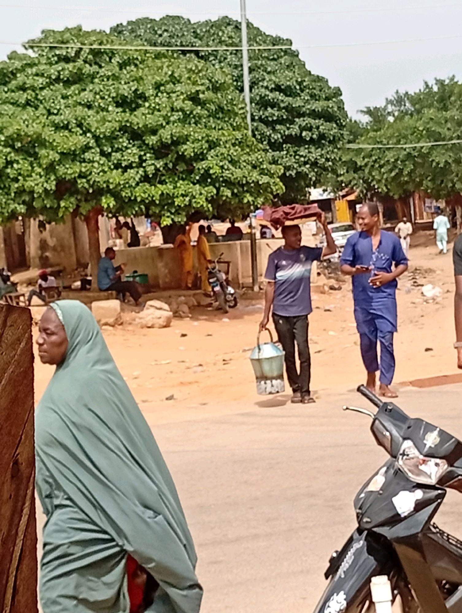 A medium shot captures a street scene in Gombe, Nigeria, under a bright sky, with lush green trees dominating the background. In the foreground, a woman wearing a pale green hijab and dress walks towards the left, her face in profile. To her right, two men walk away from the viewer, one in a dark blue t-shirt and black pants carrying a bucket and a tray on his head, the other in a blue traditional outfit, holding a phone. In the middle ground, several other individuals are visible, some sitting and others standing. A motorcycle is parked on the right side of the frame, partially obscuring the road. The ground is sandy and uneven, typical of many unpaved urban areas. Buildings and stalls are present in the background, suggesting a commercial or residential area.