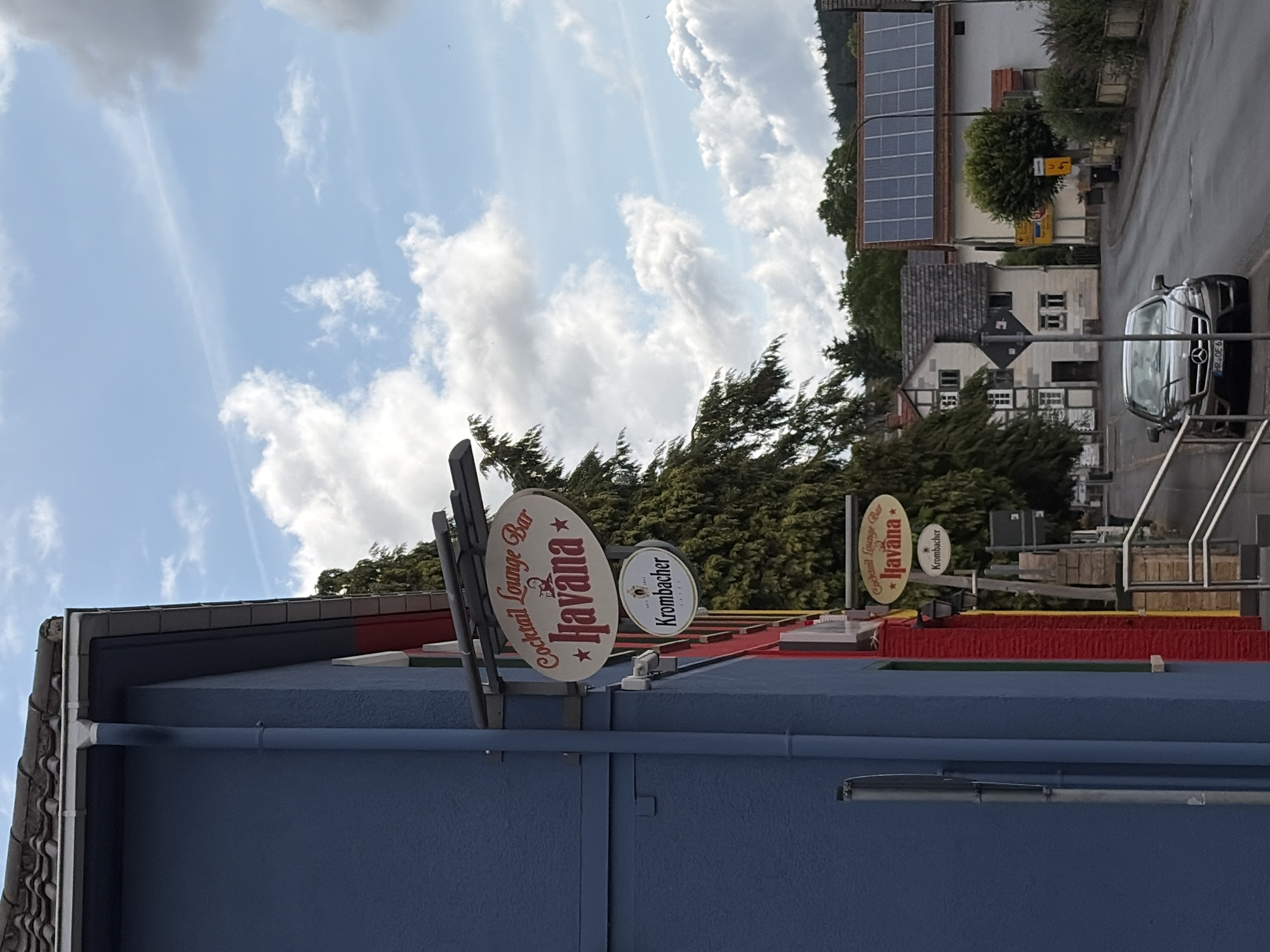 The image shows a street scene in a town. On the left side of the street is a blue building with red accents. Hanging from the building are two signs, one oval-shaped with the text "Cocktail Lounge Bar Havana" and another circular sign that says "Krombacher". Further down the street, a dark-colored sedan is parked. In the background, there are several houses, some with solar panels on their roofs, and a wooded hillside. The sky is partly cloudy.