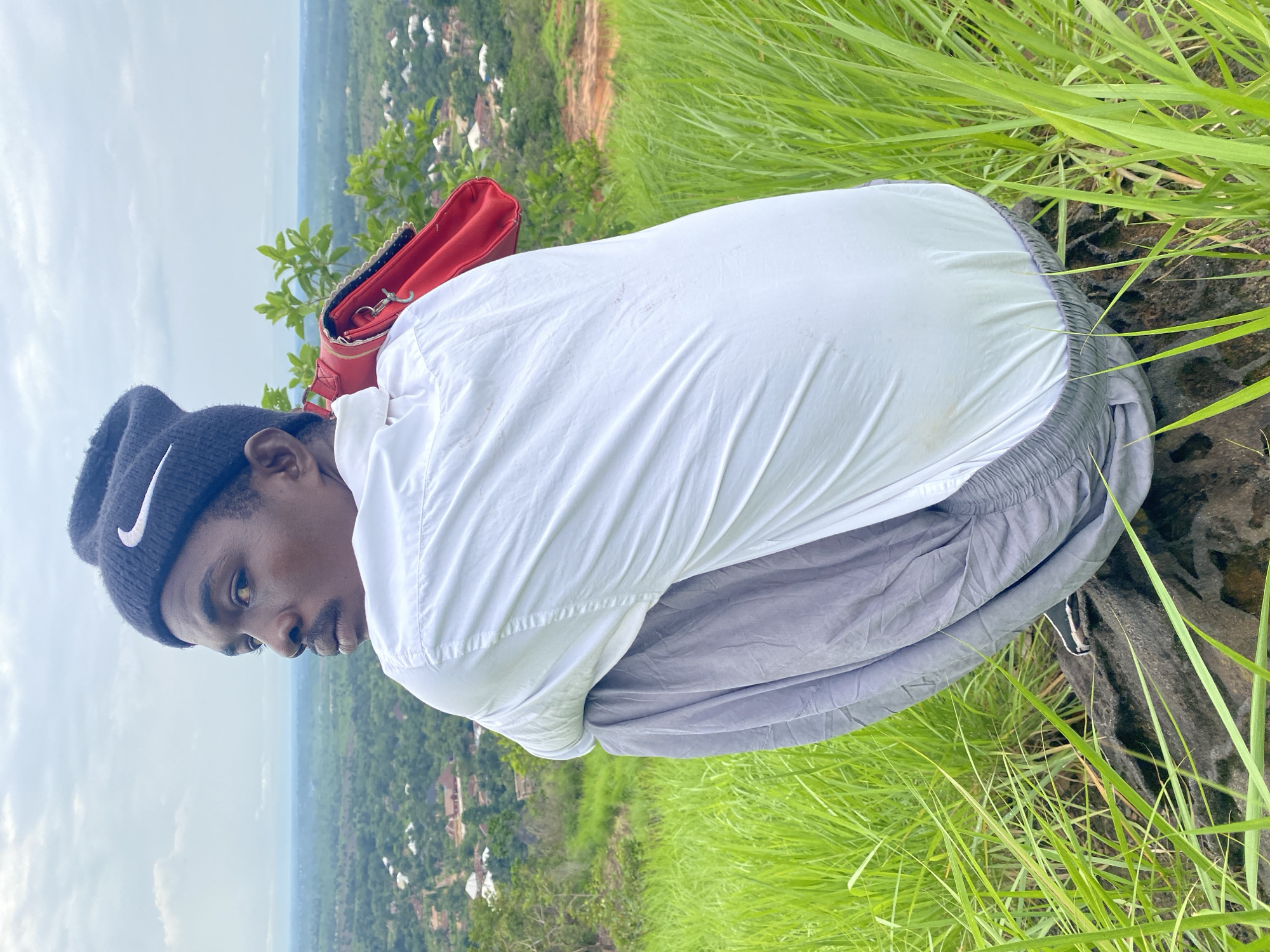 A young Black male wearing a black Nike beanie, a white button-down shirt, and gray sweatpants is crouched on a rock at the top of a hill overlooking a landscape. A bright red purse is slung over his right shoulder. The background shows a sprawling village with red-roofed houses nestled amongst lush green trees and vegetation under a cloudy sky. The foreground consists of tall, vibrant green grass. The location is identified as Ufu Esioni, Nigeria.