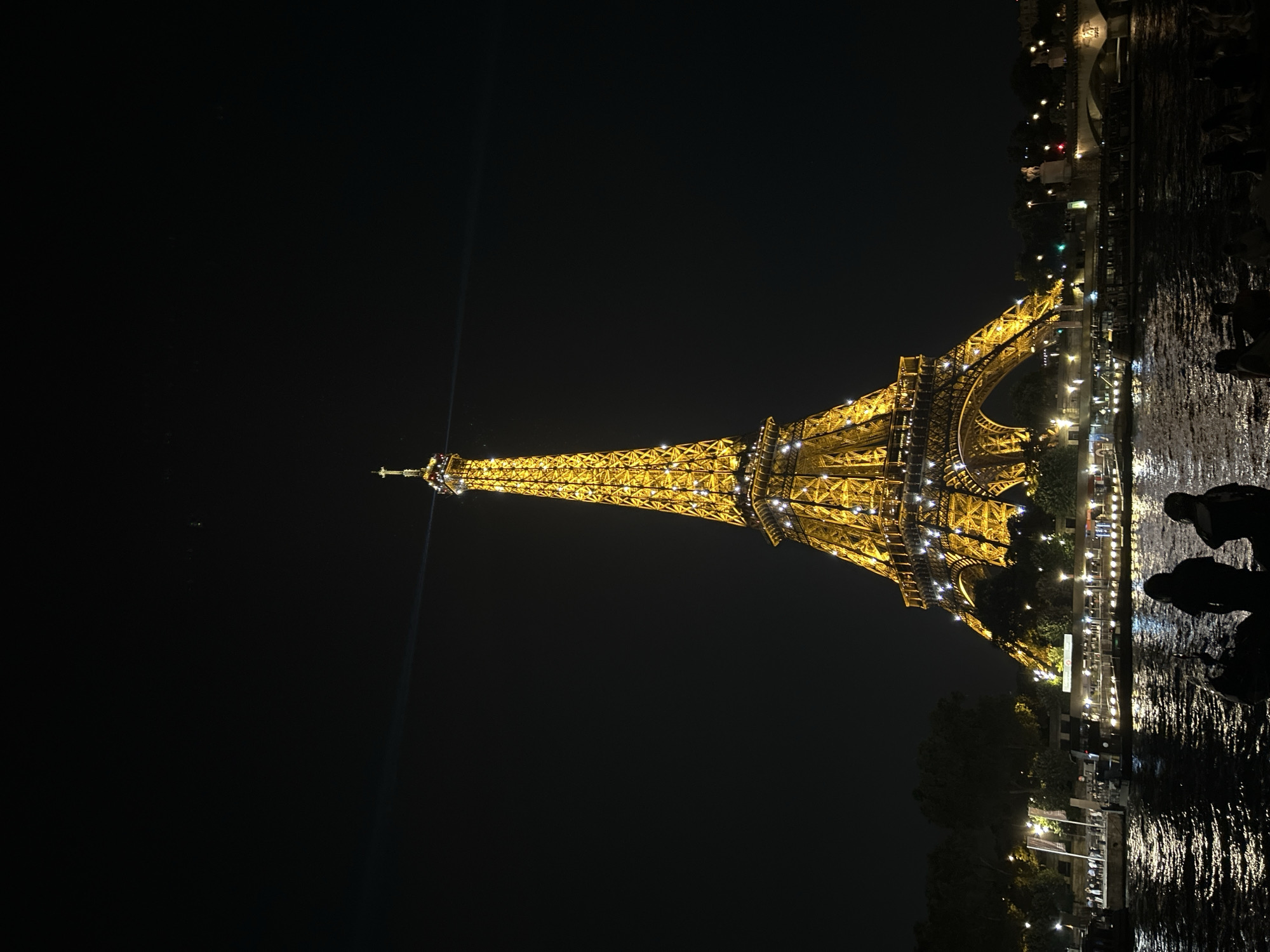 The image captures the Eiffel Tower in Paris, France, illuminated at night. The iconic landmark is a warm golden color, adorned with numerous small lights that sparkle against the dark sky. A beam of light emanates from the top of the tower, sweeping across the night. In the foreground, the dark, rippling water of a river, likely the Seine, reflects the lights of the city and the tower. Silhouetted figures of people can be seen sitting along the riverbank, gazing towards the illuminated monument. Behind the tower, the outline of trees and the distant glow of city lights are visible, along with a bridge spanning the river in the background. The scene conveys a sense of nighttime beauty and iconic Parisian ambiance.