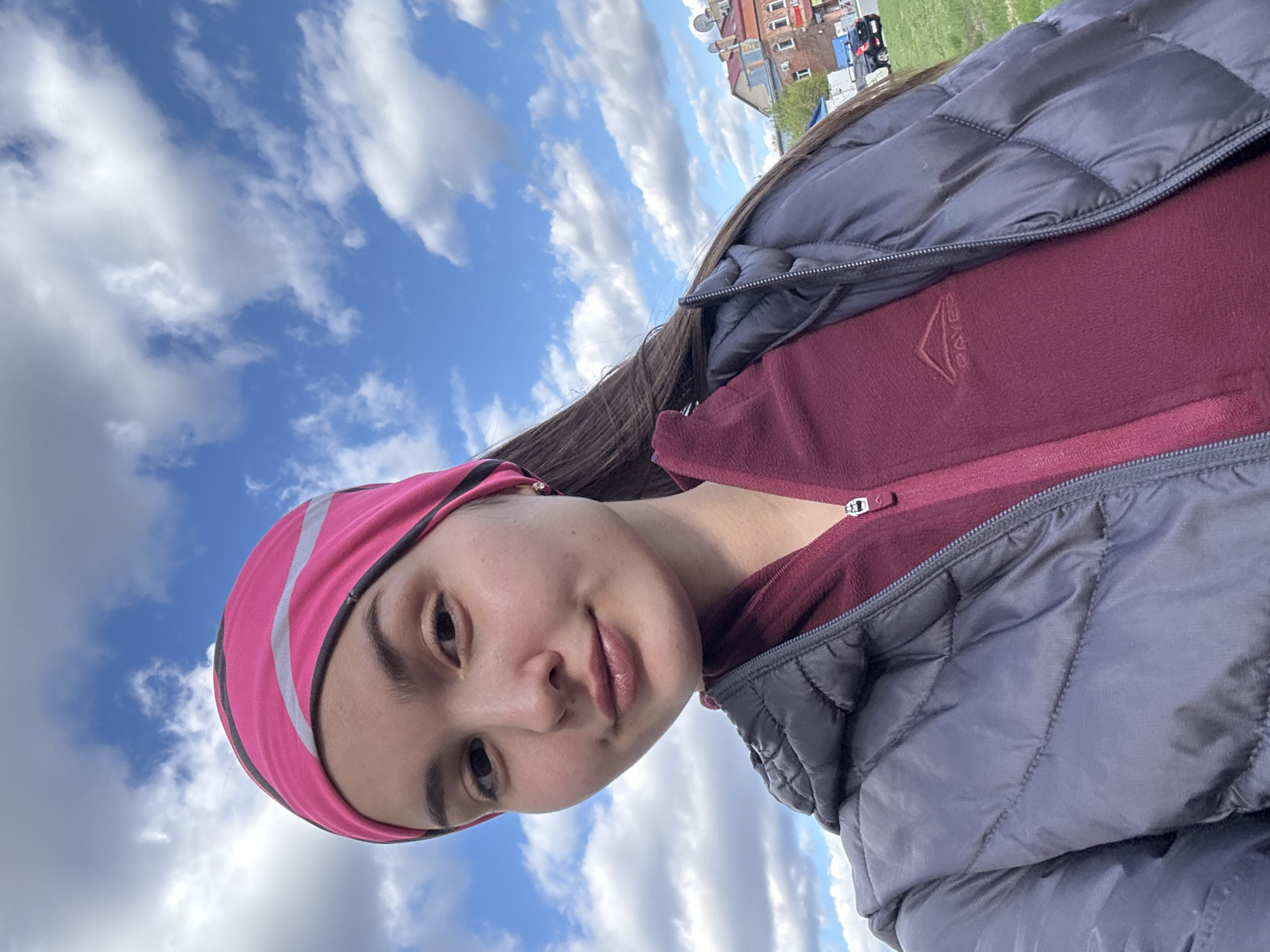A young woman wearing a pink headband, maroon fleece jacket, and gray puffer coat takes a selfie outdoors. The sky is blue with white clouds. In the distance, there are buildings and possibly vehicles, indicating a city environment. The exact location within the city of Orenburg is not determinable from the image.