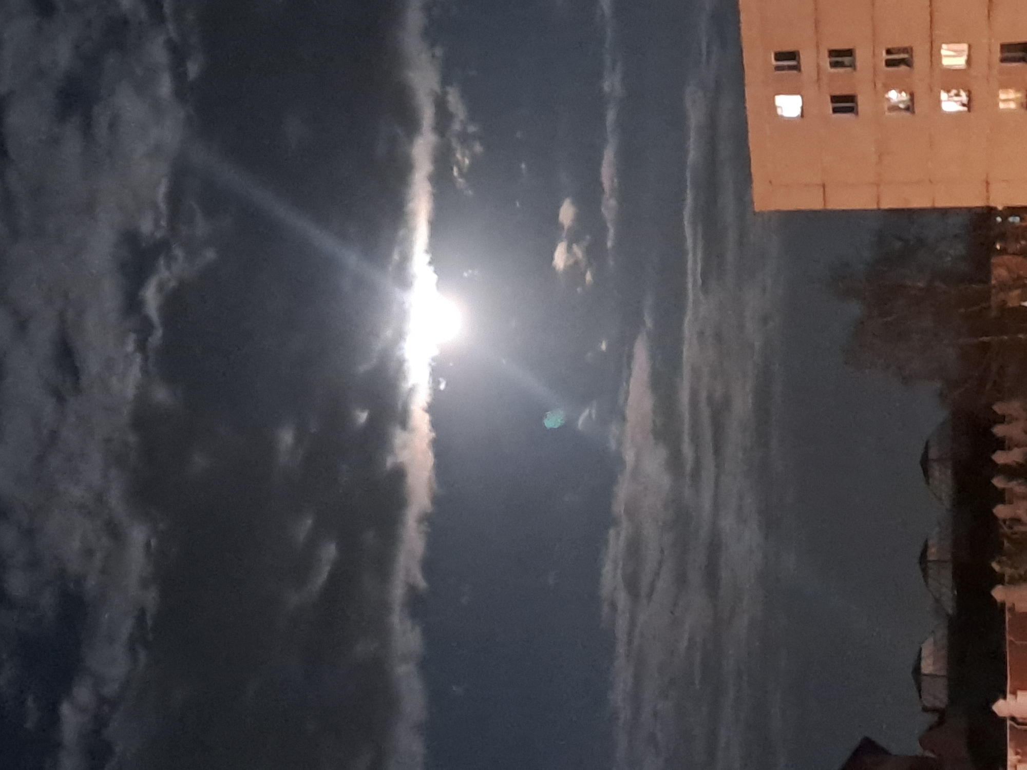 The image shows a night view in Elektrostal, Russia. The sky is predominantly dark with scattered clouds. A bright light source, possibly the moon, is visible in the center of the frame. A tall, light-colored building with multiple windows is located on the upper right of the image. Below the building, there appear to be trees and possibly structures. No people are visible in the image.
