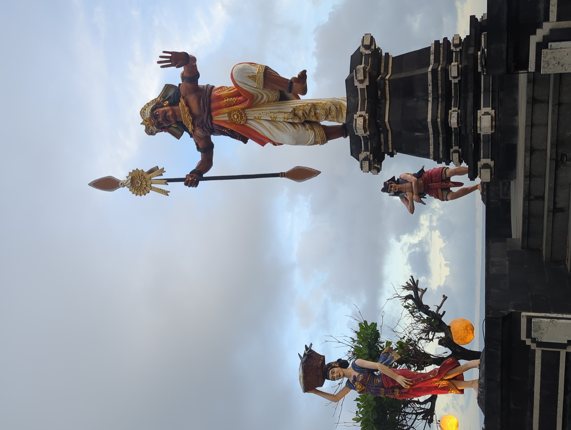The image shows a collection of statues at Taman Ayun Temple in Mengwi, Indonesia, set against a cloudy sky. The largest statue depicts a male figure holding a spear, wearing ornate attire in shades of orange, white, and gold. His right hand is raised, and he appears to be standing on a dark, multi-tiered structure. Below him, another smaller statue of a male figure is reclining. Further down and to the left, a female statue carries a basket on her head, standing near a tree adorned with glowing orange spherical lights. The statues are positioned at different levels, creating a layered visual composition.