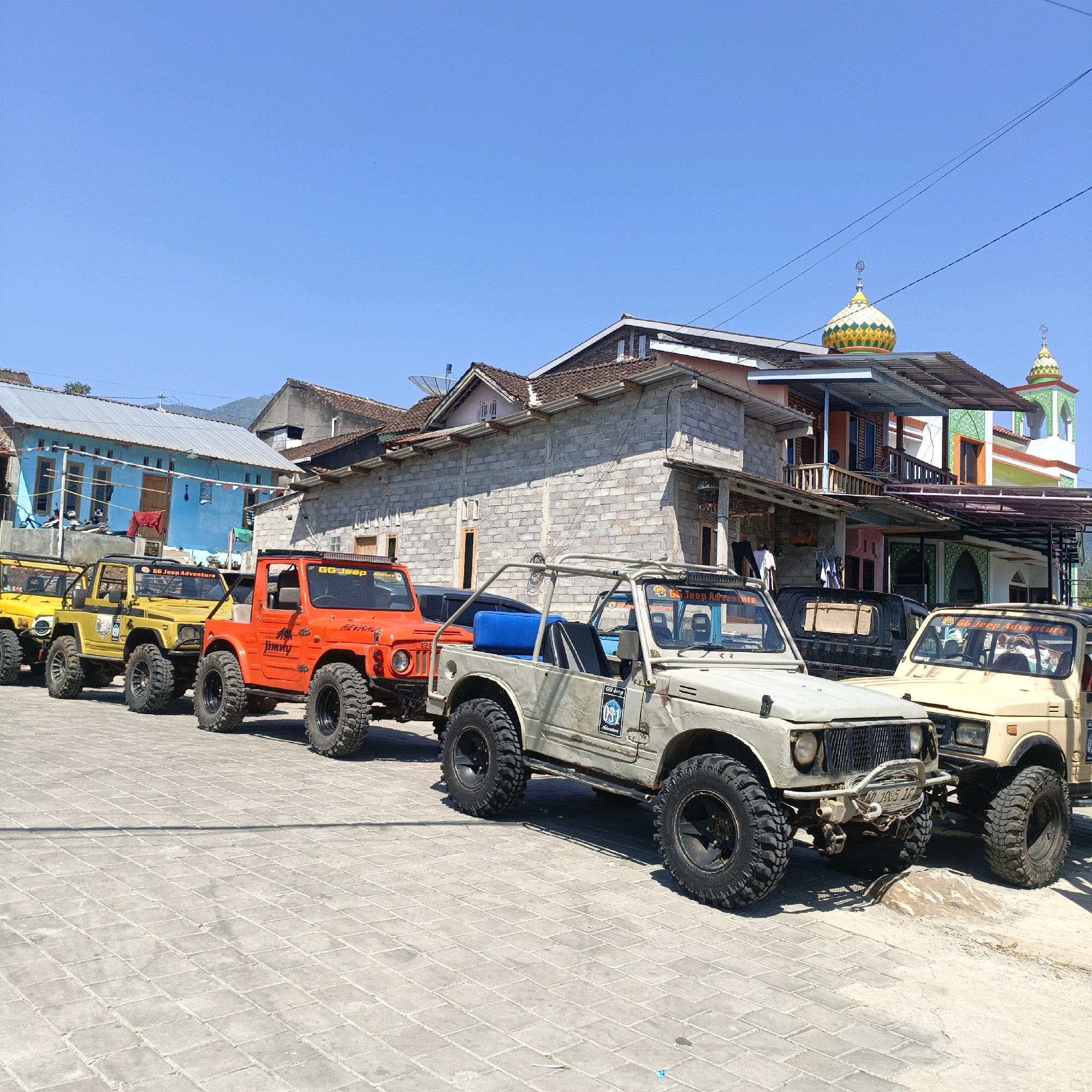In Rowoboni, Indonesia, under a clear blue sky, a line of modified off-road vehicles is parked on a paved surface. The vehicles, primarily Suzuki Jimnys, are painted in various colors including orange, yellow, and beige, and feature large, knobby tires, roll cages, and roof-mounted light bars, indicating they are prepared for off-road excursions. The vehicle in the foreground is beige and has a prominent roll cage, with a blue seat visible inside. To its left is an orange Jimny, and further down the line are yellow Jimnys. Behind the vehicles, a row of buildings is visible, including a two-story structure with a light blue facade adorned with red and white pennants. To the right, a mosque with a colorful dome and minaret stands out against the sky. The overall impression is a gathering of off-road enthusiasts in a semi-urban or rural Indonesian setting.
