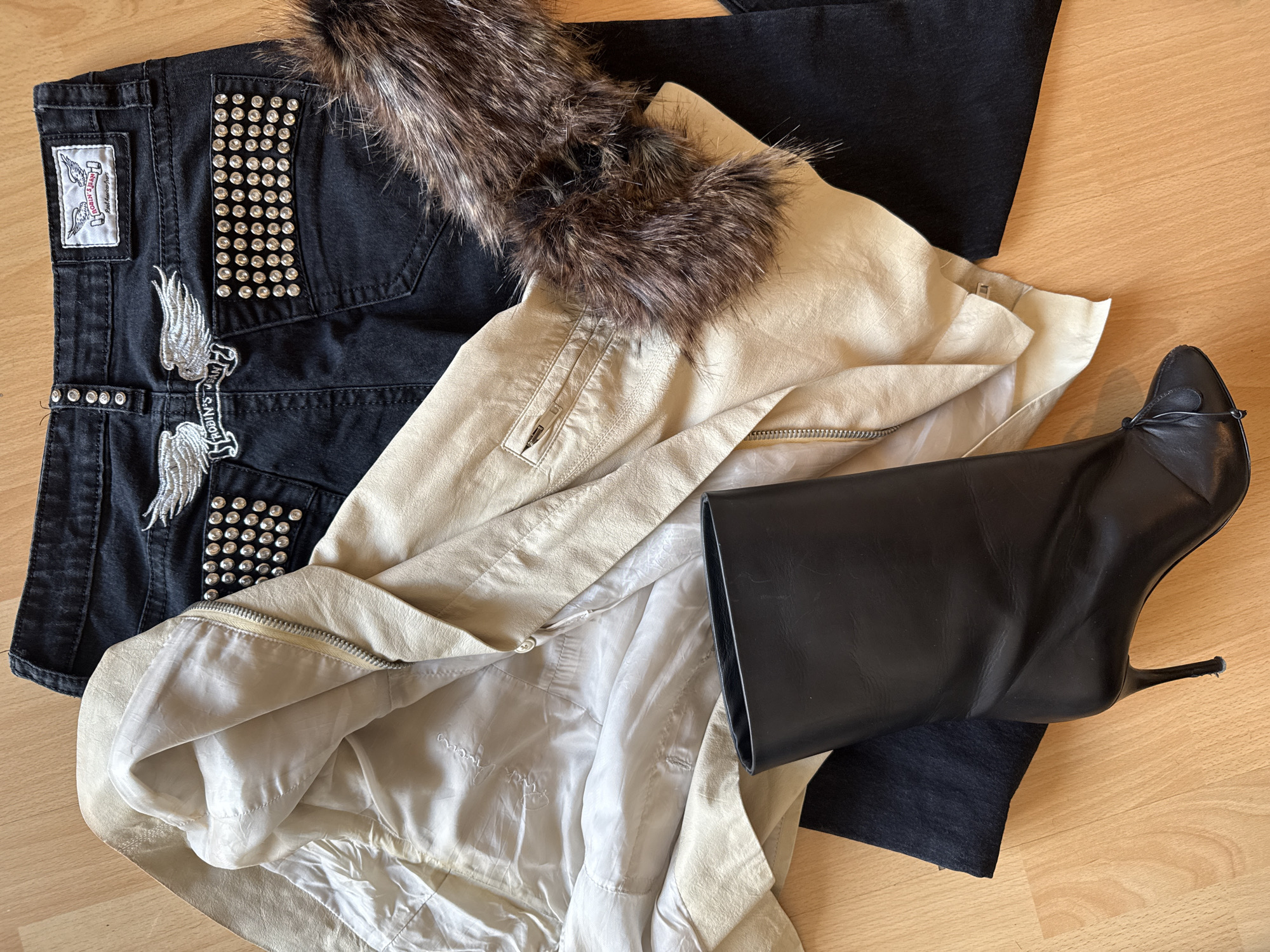 This media file presents a flat lay of fashion items arranged on a light wooden floor. The central subjects are a pair of black denim jeans, a cream-colored jacket, and a black high-heeled boot.
The black jeans are heavily embellished with silver studs covering the back pockets. They feature prominent silver embroidered winged patches, with one set on a banner that reads "ROBIN'S JEANS". A white label on the waistband also displays "ROBIN'S JEAN" with a winged logo.
Layered on top is a cream or light beige jacket, likely made of leather or faux leather, characterized by its fluffy brown faux fur collar. The jacket shows zippered details, including a pocket on the chest area and a main zipper. Its interior lining is a light, possibly satin-like, fabric.
A single black high-heeled boot is positioned on the right side of the frame. It has a sleek, pointed toe adorned with a small decorative bow or knot, and rests on a slender stiletto heel. The boot appears to be made of black leather or faux leather, with a substantial shaft suggesting a mid-calf or knee-high style.
The scene is an indoor setting, indicated by the light wooden flooring. No specific activity is occurring; the items are neatly arranged as if for display or an outfit selection. The aesthetic suggests an edgy, glamorous, or rock-chic style. No time of day, weather, or emotions are discernible.