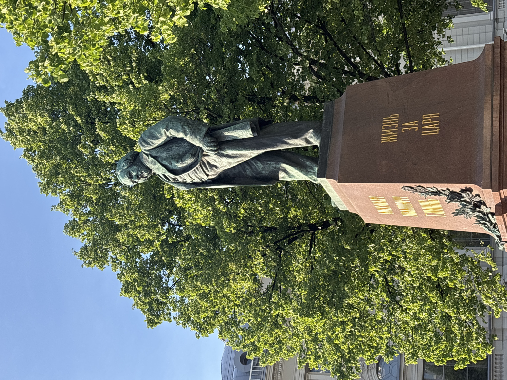 A bronze statue of a man stands on a stone pedestal in Saint Petersburg, Russia. The statue depicts a stout man with a beard, wearing a coat and trousers. He stands with his left hand on his hip and his right hand holding a folded cloth. The pedestal is made of reddish-brown stone and has inscriptions in Russian. The statue is set against a backdrop of lush green trees and a clear blue sky. In the background, a building with classical architecture is visible. The lighting suggests it is daytime, possibly late afternoon, with sunlight illuminating the scene.