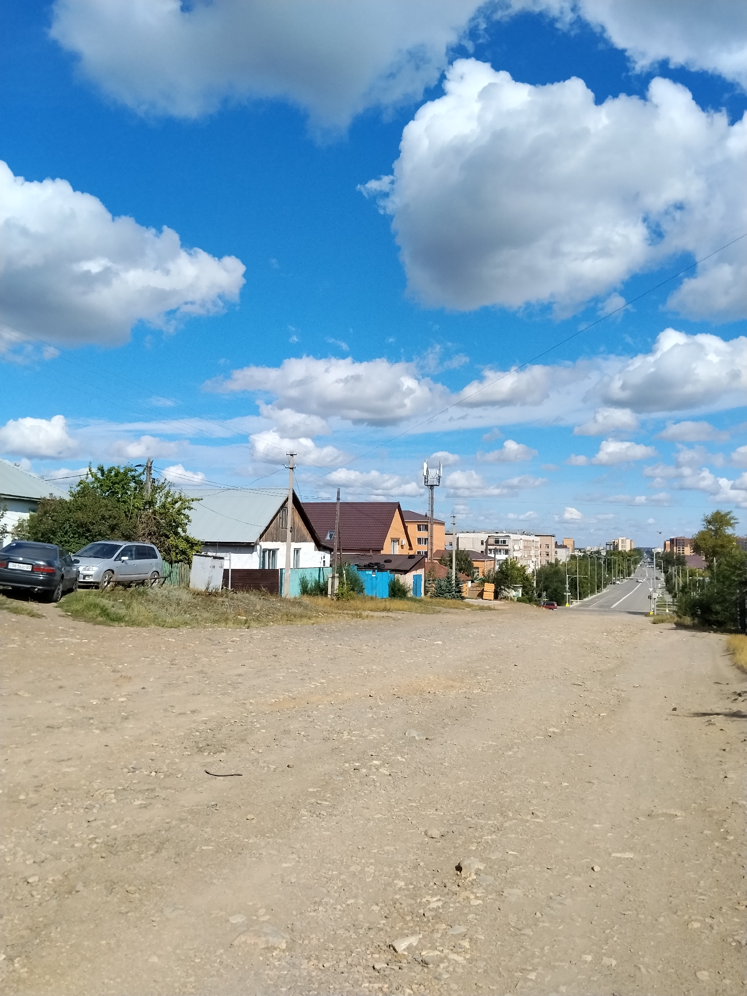 The image depicts a sunlit day in a semi-urban area, possibly on the outskirts of Kokshetau, Kazakhstan, characterized by a blend of residential houses and more developed buildings. The foreground is dominated by a dusty, unpaved dirt road with scattered pebbles, suggesting a rural or developing part of the city. To the left, two cars are parked: a dark-colored sedan and a silver minivan. Behind the cars, a low fence borders a property with a modest house featuring a light-colored facade and a dark roof. Further to the right, a road stretches into the distance, lined with trees and a mix of buildings, including several multi-story structures that indicate a more urbanized area. A cell tower is visible in the middle ground, adding to the modern infrastructure in the scene. The sky is a vibrant blue, dotted with fluffy white clouds, contributing to a clear and bright atmosphere. The overall impression is of a transitional zone within a city, where rural elements meet urban development.