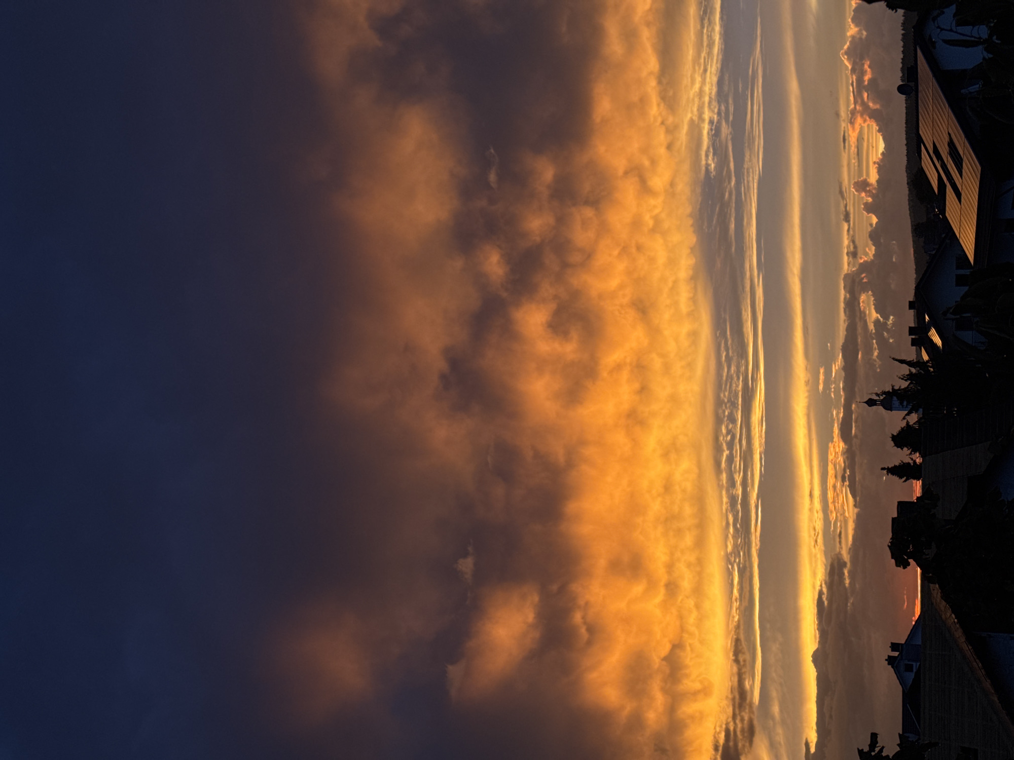 This is a photograph of a sunset over the town of Wettstetten, Germany. The sky is filled with dramatic clouds, illuminated in vibrant shades of orange and yellow, contrasting with the deep purple and blue of the upper sky. Silhouetted against the colorful sky are rooftops, trees, and a church steeple, indicating a residential area. The scene captures the transition from day to night, with the last rays of sunlight painting the clouds.