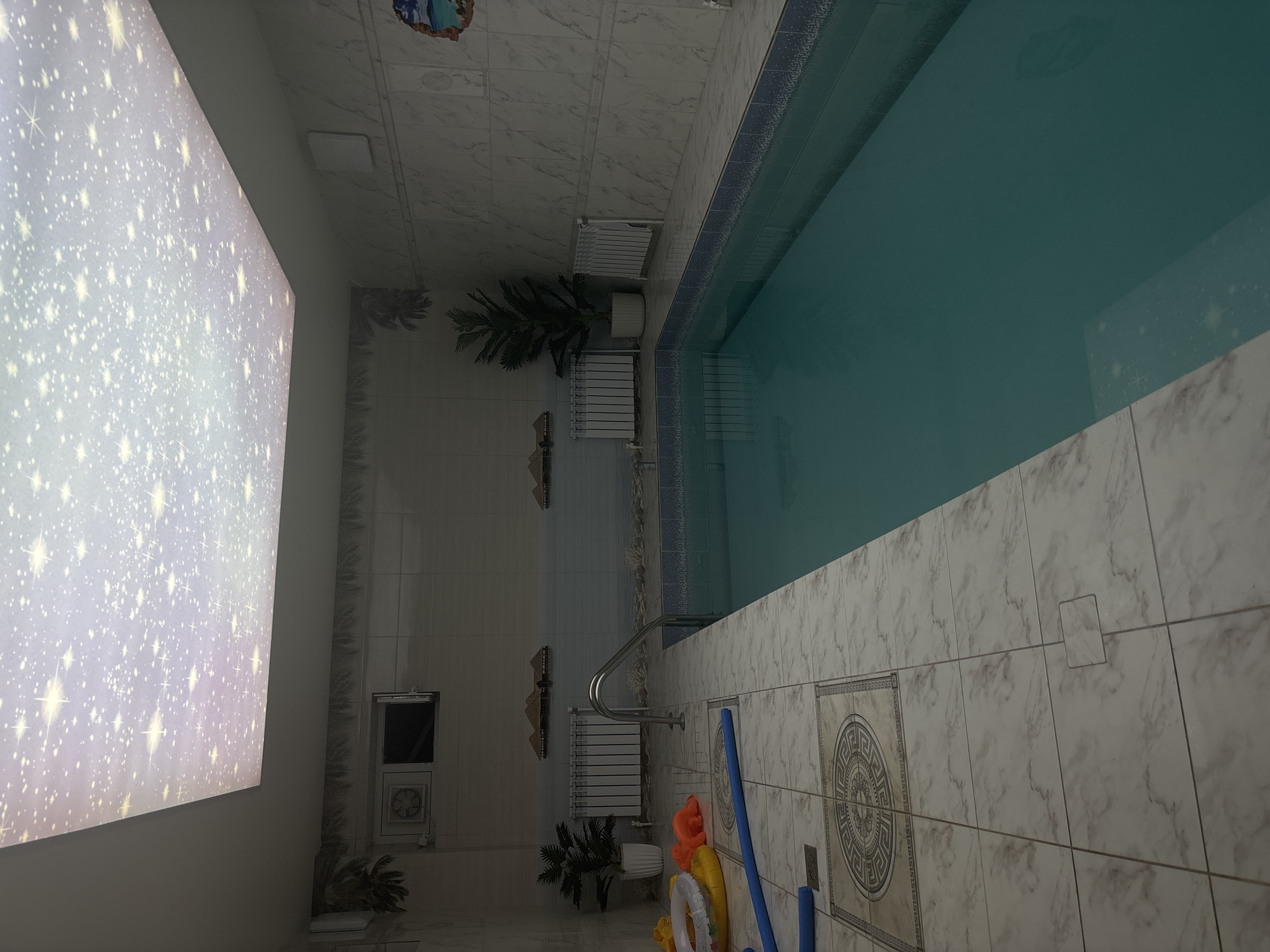 The image shows an indoor swimming pool in a room tiled with white marble-patterned tiles. A narrow pool with turquoise water runs along the right side of the image. There are heating radiators installed along the wall adjacent to the pool, as well as potted plants. In the lower part of the frame, there are pool noodles and inflatable pool toys. The ceiling has a starry sky design. The location is identified as being in Chaikovsky, Russia, based on a forensic analysis of patterns matching known building and architectural details in the city.
