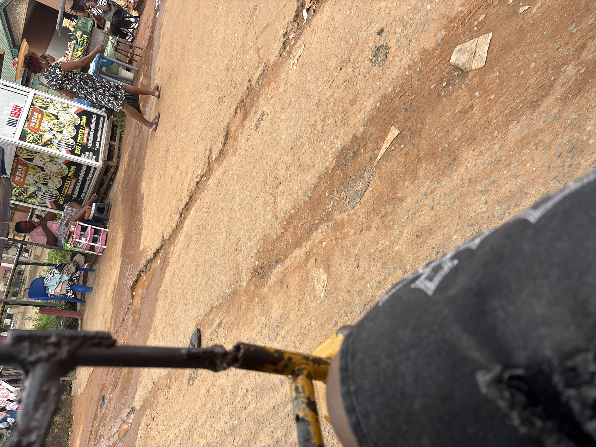 The image depicts an outdoor scene, likely on a street in Nkpor, Nigeria. The foreground is dominated by a portion of dark, possibly denim, trousers with visible distressed areas and what appear to be white graphic designs. A weathered, dark metal object, part of a vehicle or structure, occupies the left side of the frame.
The midground shows a dirt and gravel road with noticeable erosion and muddy patches, indicating recent rainfall or poor drainage. Scattered debris, including paper fragments, is present on the ground.
In the background, several individuals are visible. On the left, a person in a pink shirt sits on a blue plastic chair, holding a phone. Next to them is a small, pink, tiered plastic shelf holding various items of clothing. Further to the right, a woman in a patterned dress walks away from the camera. Behind her, a stand with advertising posters, including one for "JELE GALAXY" and "10 STAR SHARWARMA," is visible. To the far right, a person in a striped shirt and dark trousers sits on a chair, looking towards the left. A green stall with produce, possibly bananas, is also present in the background. The overall environment suggests a bustling roadside area with commercial activity.