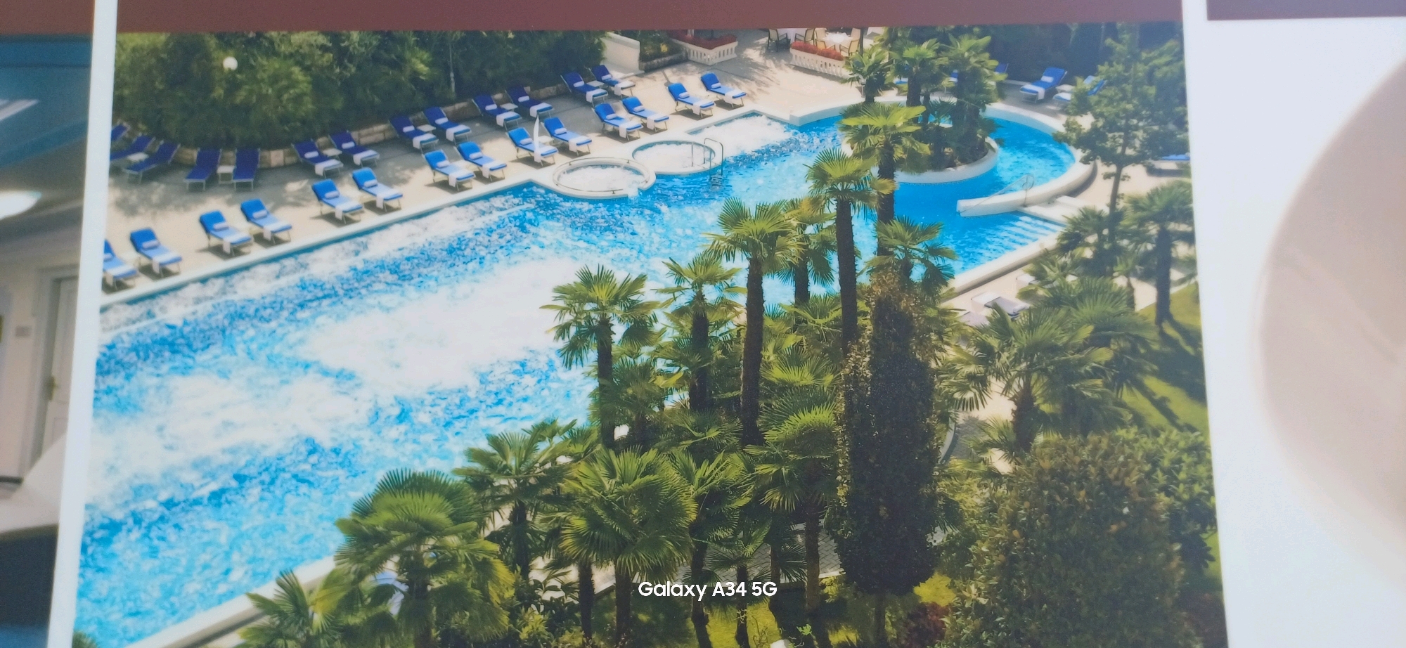 This is an aerial view of a resort with a large, irregularly shaped swimming pool and a separate circular jacuzzi area. The pool is surrounded by numerous blue lounge chairs, many of which are empty. Lush green palm trees and other foliage are scattered throughout the resort grounds, casting shadows on the pavement and the water. In the background, there are buildings with red roofs and outdoor seating areas with tables and chairs. The image appears to be a promotional photograph, as indicated by the watermark "Galaxy A34 5G" in the lower right corner. This scene is characteristic of a luxury hotel in Abano Terme, Italy, a city renowned for its thermal spas and resorts.
