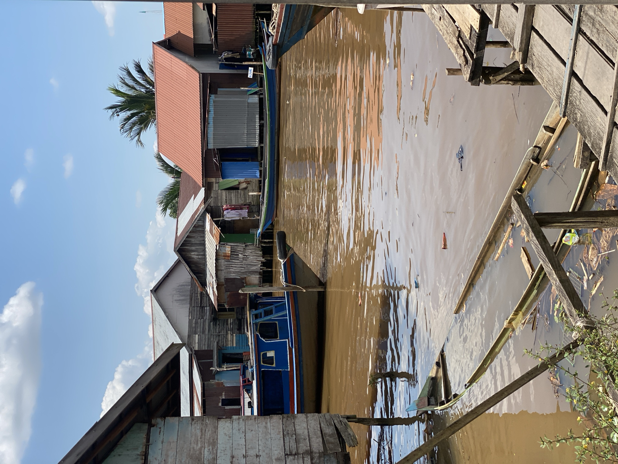 This image depicts a view of the village of Berangas Timur, Indonesia, centered on a murky brown waterway. Wooden stilted houses with corrugated metal and wooden siding line the water's edge, with some showing signs of disrepair. Several boats are visible in the water. In the foreground, a partially submerged wooden boat rests at an angle, filled with water and debris. A larger blue and orange boat is moored in the mid-ground. The sky is blue with scattered white clouds, and a palm tree is visible behind the houses. The overall scene conveys a sense of a rural, water-based community.