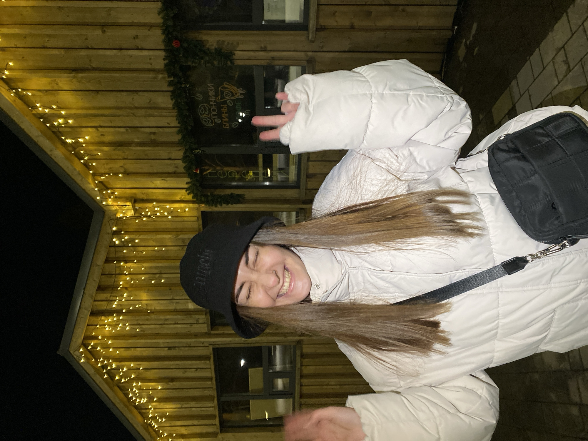 The image shows a woman in a white puffer jacket and a black hat with Cyrillic writing standing outside a wooden building decorated with string lights and a sign in Russian. The woman is smiling and holding up two fingers in a "peace" sign. She is wearing a black shoulder bag with a black strap. The building appears to be a cafe or restaurant, based on the writing on the sign. The photo was taken at night. Based on the Cyrillic writing and the prompt's direction, the location is presumably in Votkinsk, Russia.