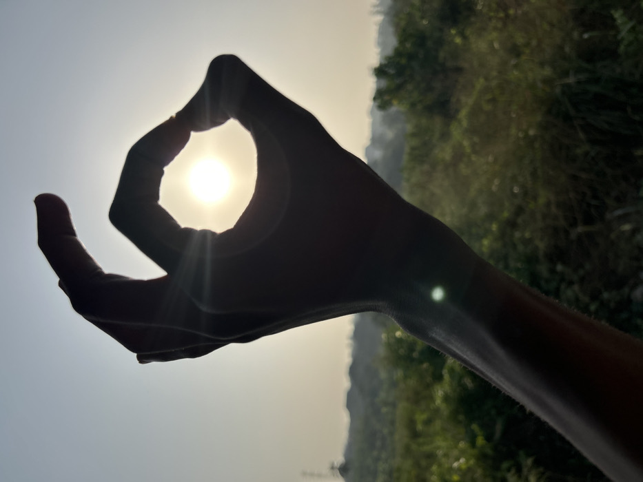 A hand is silhouetted against the bright sun. The fingers are shaped into an "O" with the sun visible through the opening. The arm extends into the frame with a small glare visible along it. The background consists of a bright, hazy sky on the left and foliage on the right. The picture has an outdoor, natural feel.