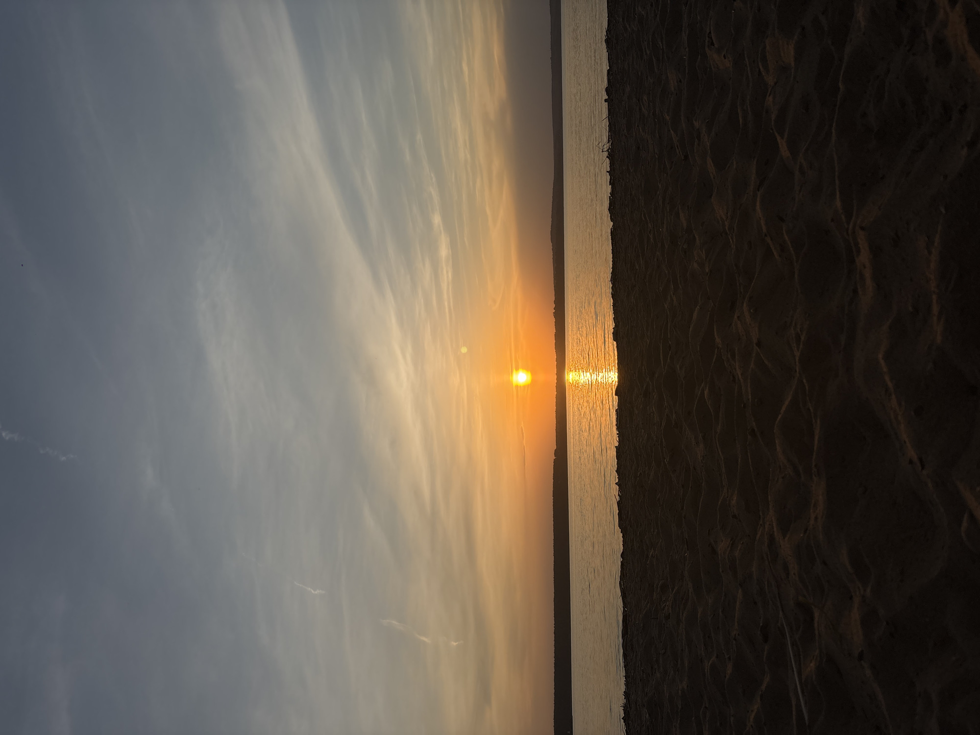 The image shows a sunset over a body of water, likely a river or lake, in Perm, Russia. The sun is positioned near the horizon, reflecting brightly on the water's surface. The sky has light gray clouds. The shoreline consists of sandy terrain.