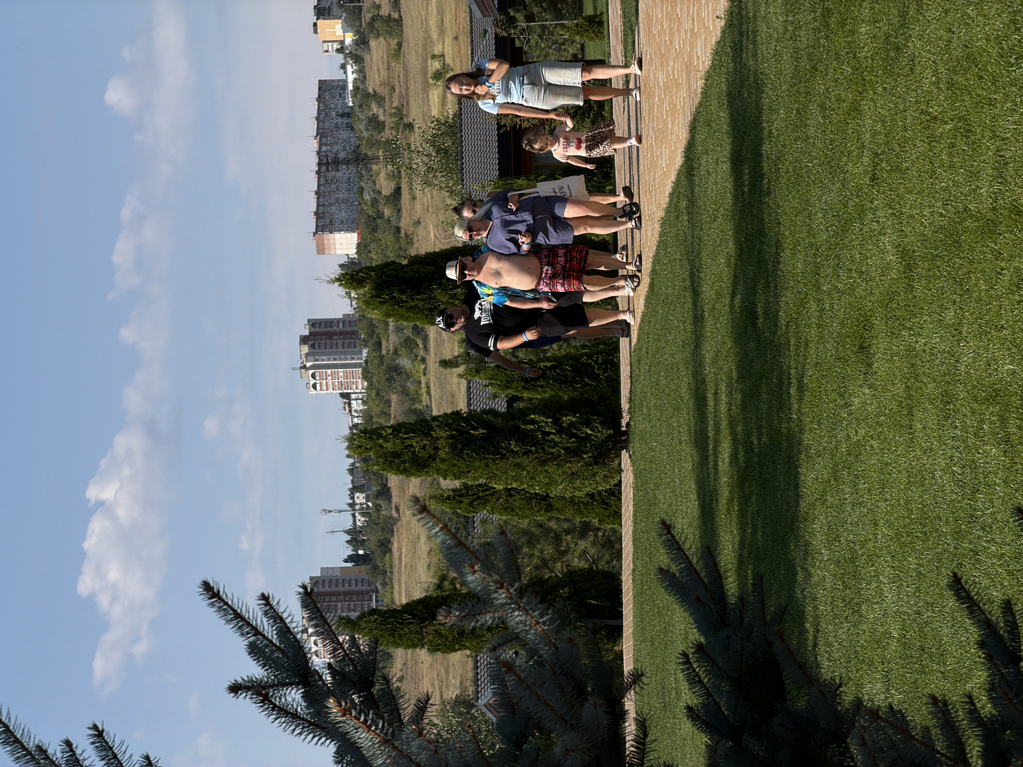 In Kamyshin, Russia, on a bright, sunny day, a group of seven people walks along a paved pathway in a park-like setting. The pathway is constructed of light-colored bricks laid in a herringbone pattern. To the left, a meticulously manicured lawn is visible, with a portion of a dark green evergreen tree's branches in the foreground. Behind the pathway and to the right, there are buildings with dark tiled roofs and several tall, dark green evergreen trees. In the background, a landscape of rolling hills is dotted with more buildings, including apartment complexes and what appears to be a cell tower. The sky is a clear blue with scattered white clouds.
The group consists of five adults and two children. Starting from the left, a man wearing a black t-shirt, shorts, and a black baseball cap walks with sunglasses on. Next to him is a man in a brightly colored, tropical-patterned shirt and patterned shorts, wearing a straw hat. Following him is a woman in a navy blue jumpsuit, holding a white shopping bag with black lettering. Behind her, a woman with blonde hair is partially visible. Further to the right, a woman in a light blue t-shirt and denim shorts walks with a young girl holding her hand. The girl wears a patterned dress with a cherry print and is walking in the same direction as the adults. The group appears to be casually strolling through the park.