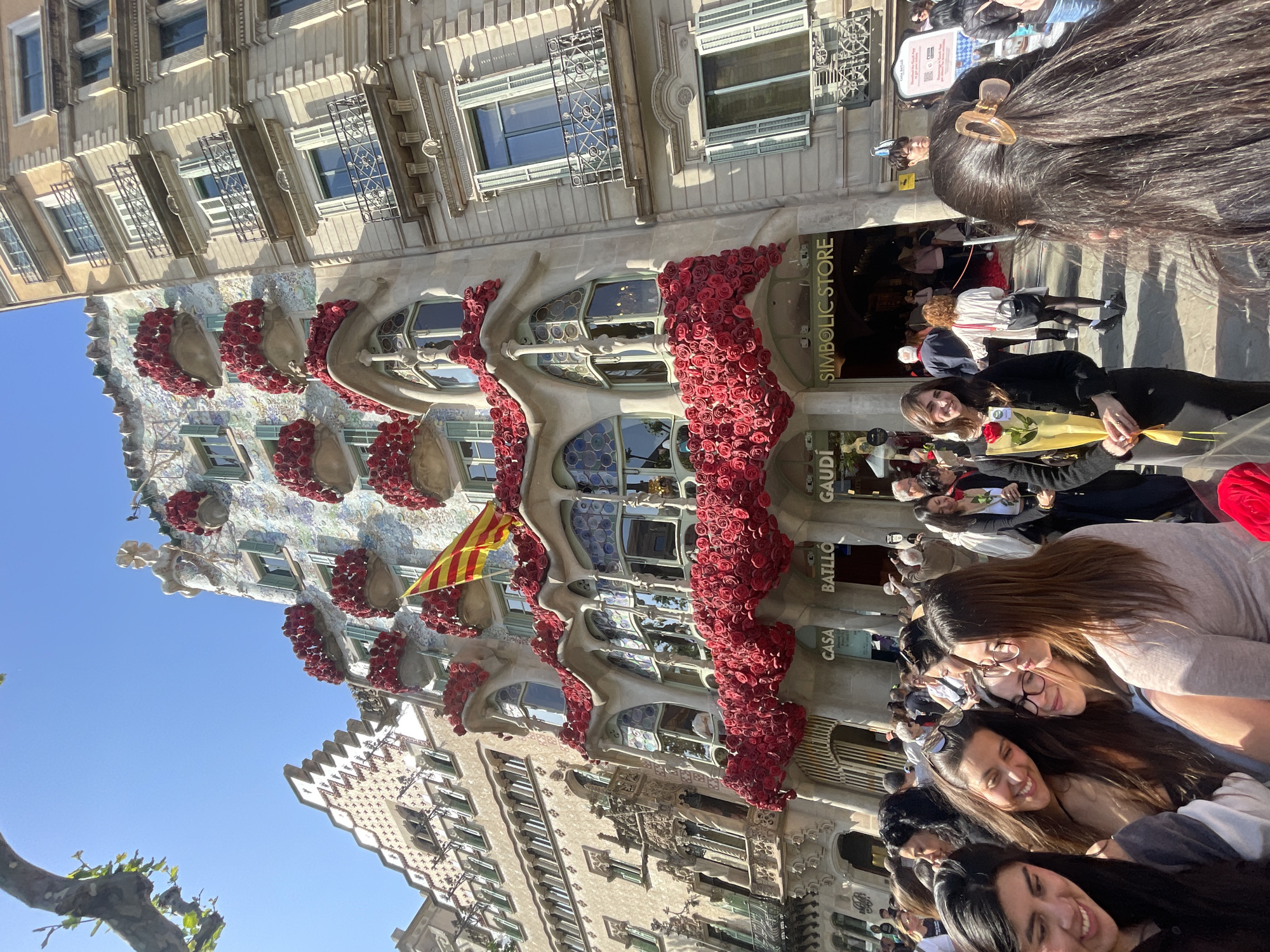An upright, sunlit street scene captures a vibrant crowd in front of Casa Batlló in Barcelona, Spain, adorned extensively with red roses. The unique modernist facade of the building, designed by Antoni Gaudí, features organic shapes, skull-like balconies, and intricate tilework. A yellow and red striped Catalan flag (Senyera) hangs from one of the upper balconies.
Numerous people, predominantly women, are visible in the foreground and mid-ground. In the immediate foreground, the backs of several heads with long dark hair are prominent, suggesting they are facing the building. Further back, a woman with dark hair is holding a single red rose, smiling towards the camera. Other individuals are seen walking or standing, observing the decorated landmark. Many faces show smiles and a cheerful demeanor, indicative of a festive atmosphere.
The extensive red rose decoration on Casa Batlló strongly suggests the celebration of Sant Jordi's Day (Saint George's Day), a major cultural festival in Catalonia where roses and books are exchanged. The bright, clear blue sky above and the strong sunlight indicate a daytime setting.
Visible text on storefront signs includes "SIMBOLIC STORE," and partially visible text on the building itself reads "GAUDÍ," "BATLLÓ," and "CASA," confirming the iconic location. The overall scene portrays a bustling, celebratory event at a renowned architectural site.