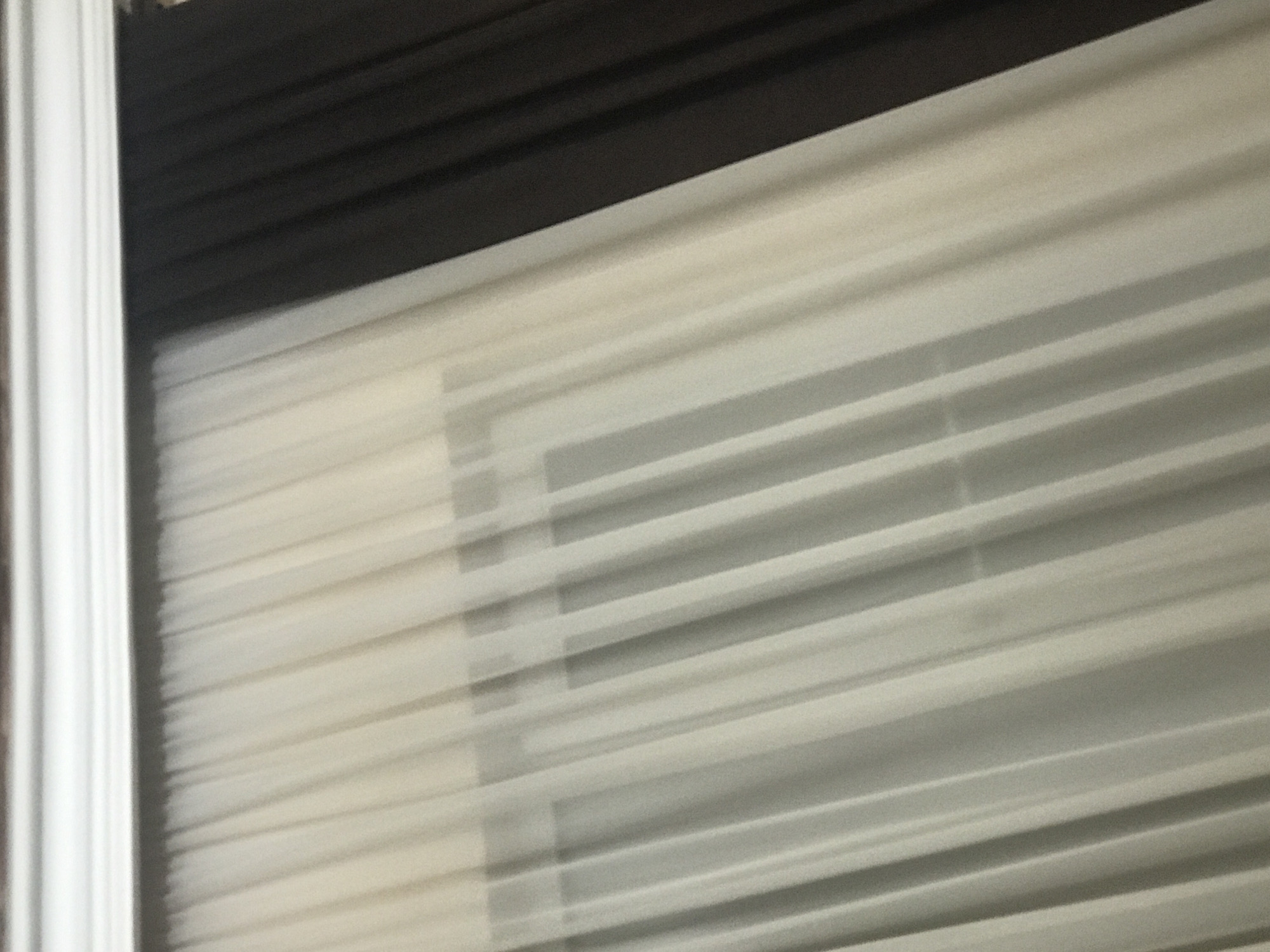 A close-up, slightly blurred image showing partially closed, light-colored window blinds. The slats are horizontal, with a dark header and a white vertical frame visible on the left. The location is not identifiable.