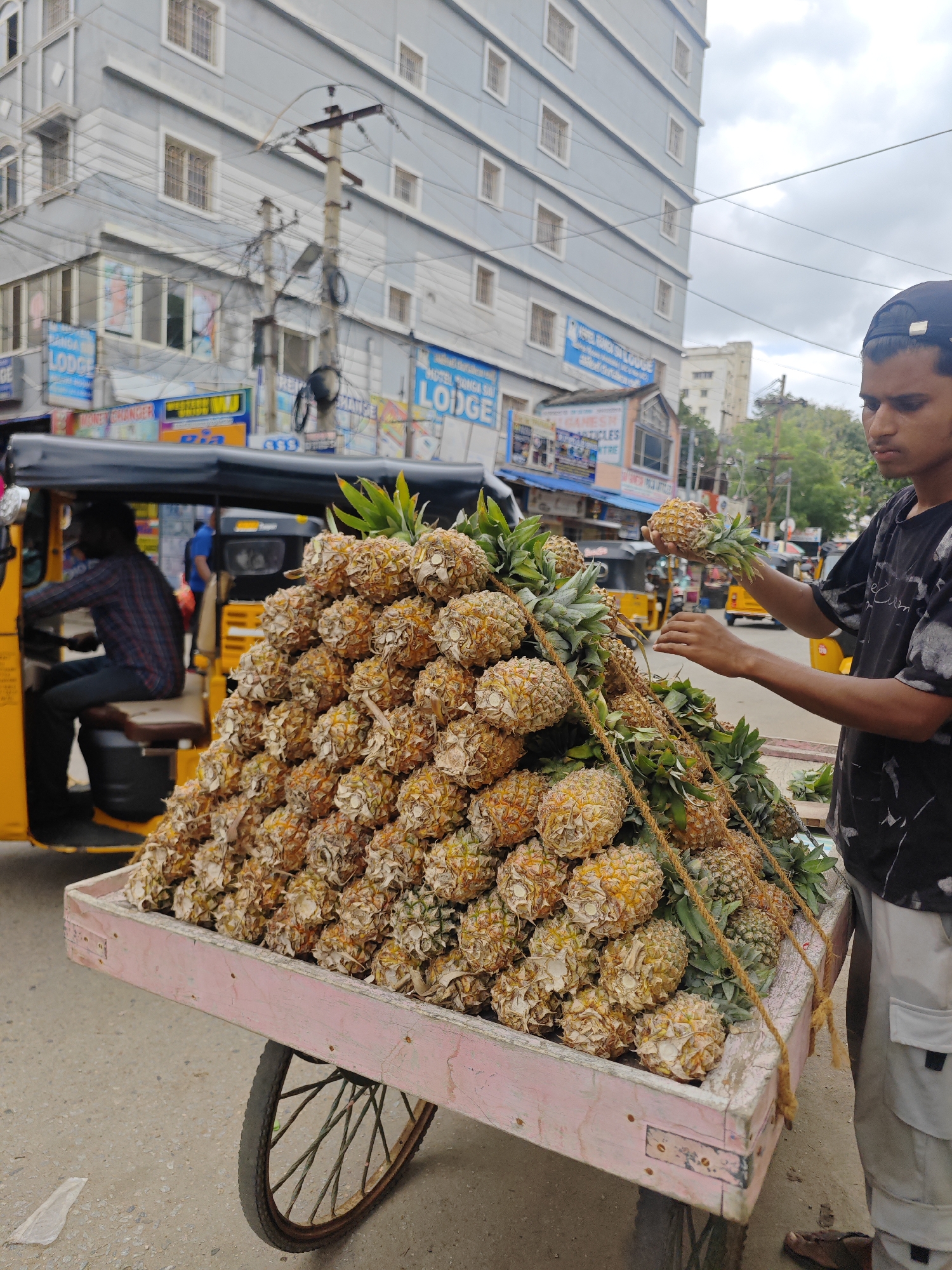 In Puttaparthi, India, a young man wearing a dark t-shirt and a baseball cap stands next to a cart laden with a large pile of pineapples. He is holding one pineapple in his right hand and reaching with his left, seemingly to select another or arrange the display. The cart, a rustic wooden structure painted pink and showing signs of wear, has a single wheel visible. Behind the cart, an auto-rickshaw is parked, with a driver visible in the driver's seat. The background reveals a busy street scene with a multi-story building adorned with signage, including what appears to be "LODGE" in English. Electrical wires crisscross the sky, and other vehicles and pedestrians are visible in the distance, suggesting an active urban environment. The overall impression is of a vibrant street market scene.