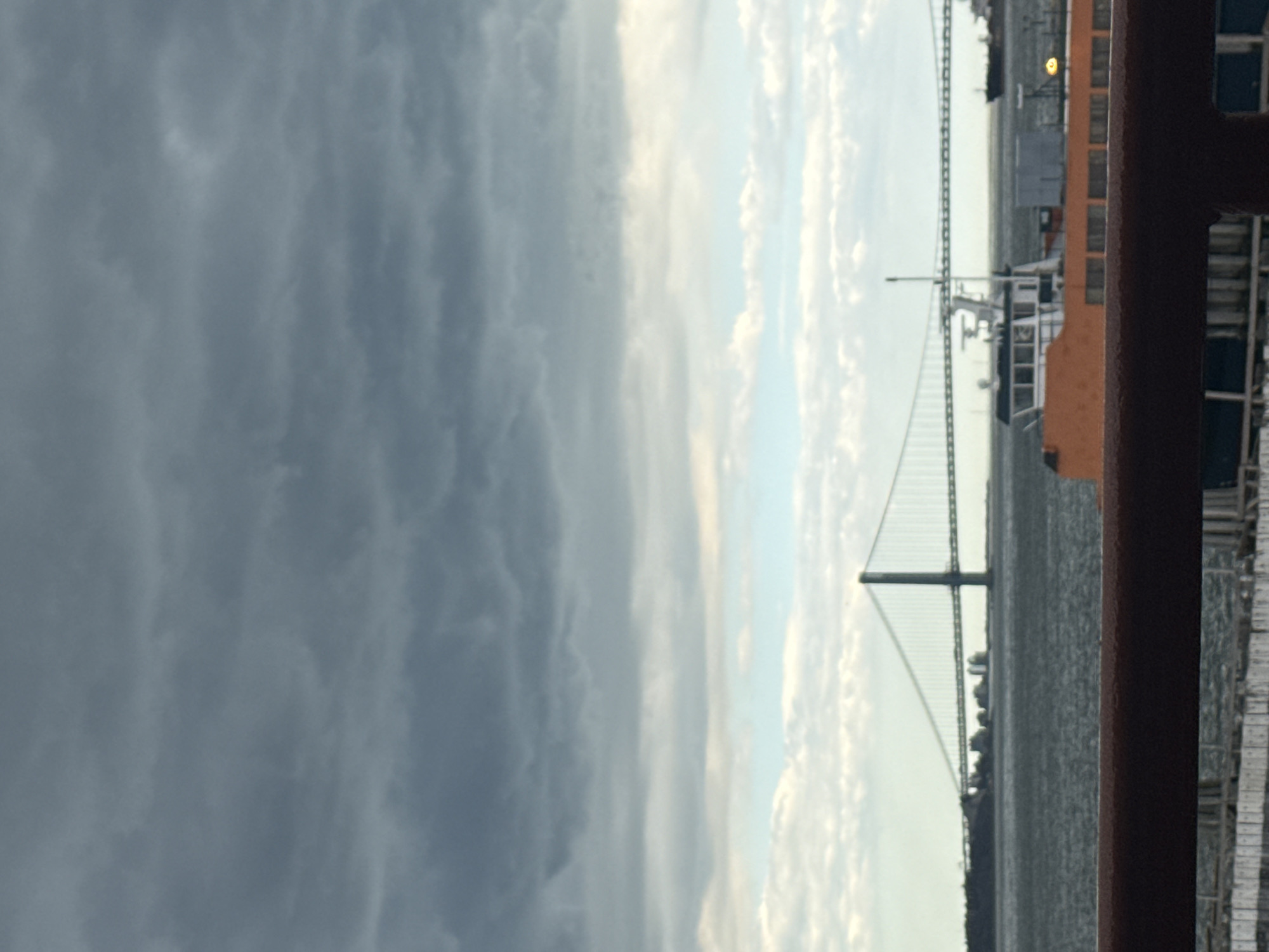 The image captures a view of the Verrazzano-Narrows Bridge in New York City from a boat or ferry. The bridge spans the Narrows strait, connecting Staten Island and Brooklyn. A large ferry is visible, positioned near the bridge. The sky is partly cloudy with storm clouds in the background, indicating a potentially overcast day. The photograph is taken from behind a reddish-brown railing, suggesting the perspective of a passenger on the vessel.