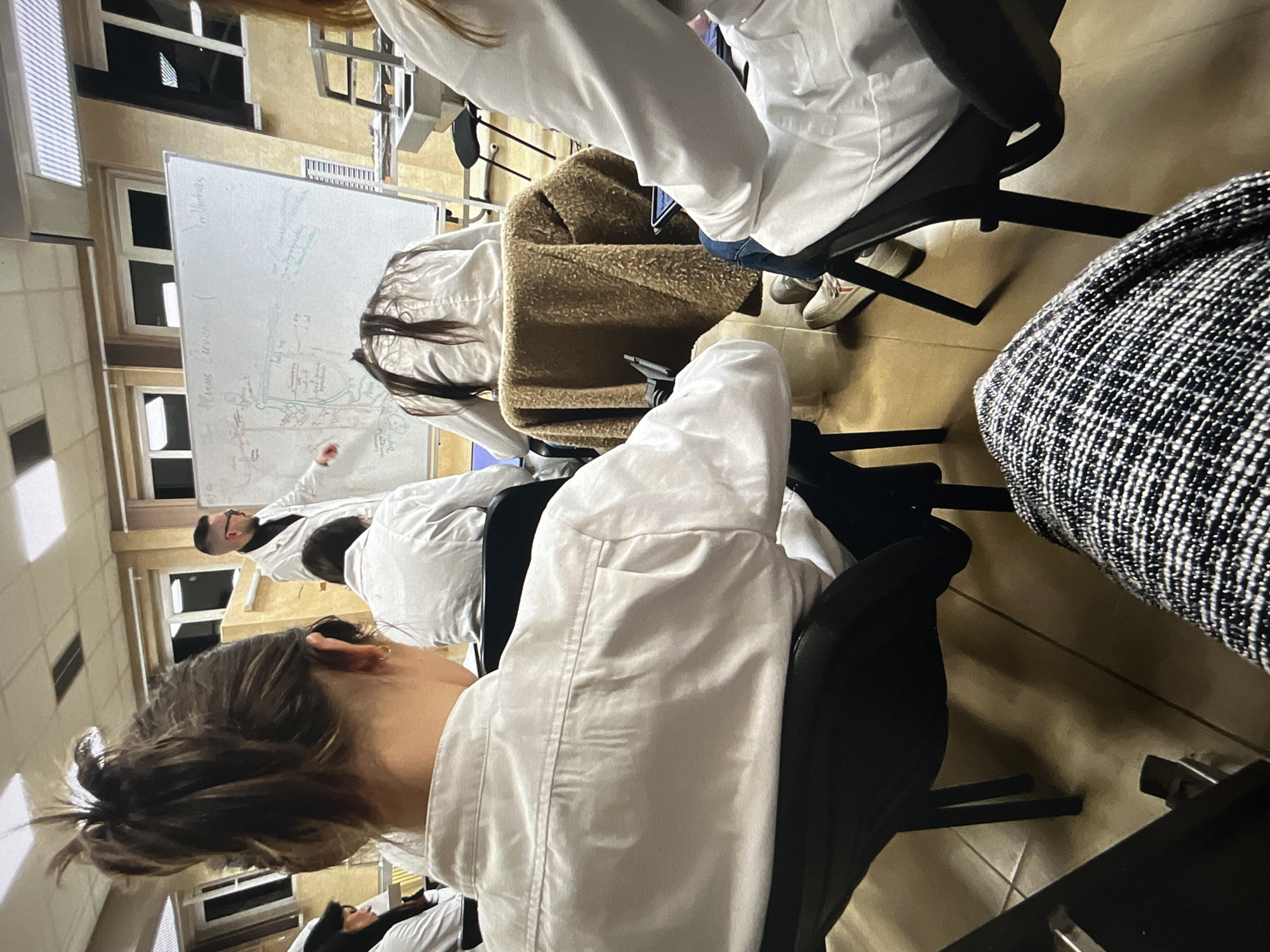 The image shows a classroom setting, possibly a lecture or presentation. Several individuals are seated in black chairs, most of them wearing white lab coats, suggesting a science or medical environment. A person, presumably the instructor, is standing near a whiteboard on the left side of the image, possibly explaining a diagram or notes written on it. The room has tile flooring, and the lighting appears to be artificial. The location is unknown.