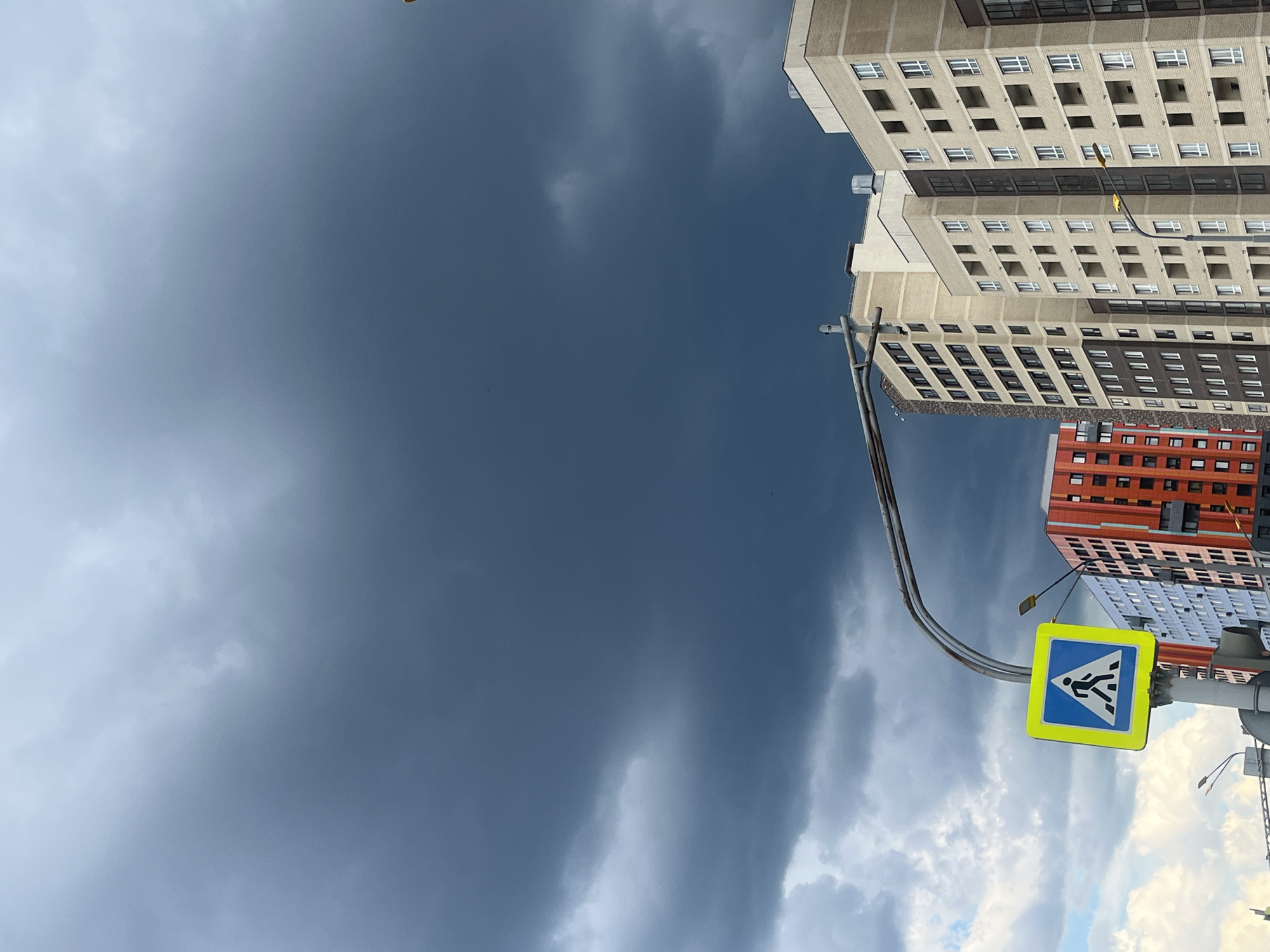 The image shows a street scene in Yekaterinburg, Russia, with a dark, cloudy sky overhead. In the foreground, a pedestrian crossing sign is visible on a pole. Behind the sign, several multi-story buildings are present, some of which appear to be under construction or newly built. The buildings vary in color and architectural style. The sky is filled with heavy, dark clouds, suggesting impending rain or a storm. The overall atmosphere is somewhat dramatic due to the contrast between the urban landscape and the ominous sky.