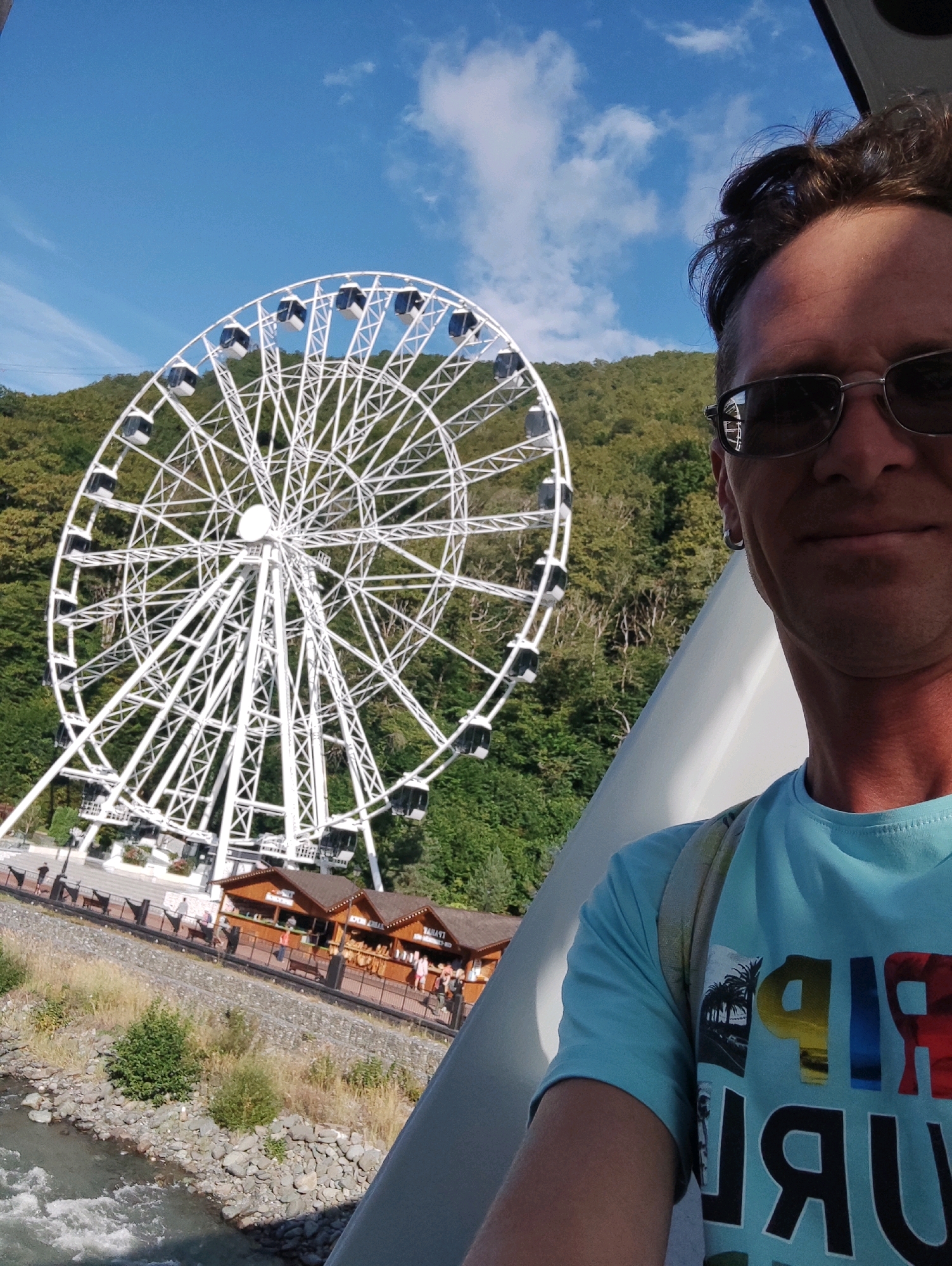 In Estosadok, Russia, a man with sunglasses and a blue t-shirt takes a selfie in front of a large white Ferris wheel. The Ferris wheel is set against a backdrop of lush green trees on a hillside and a bright blue sky with scattered clouds. The man's t-shirt has a colorful graphic with the word "RIP" and other illegible text. In the background, there are several small wooden structures that appear to be shops or food stalls, and a river flows alongside the rocky bank.