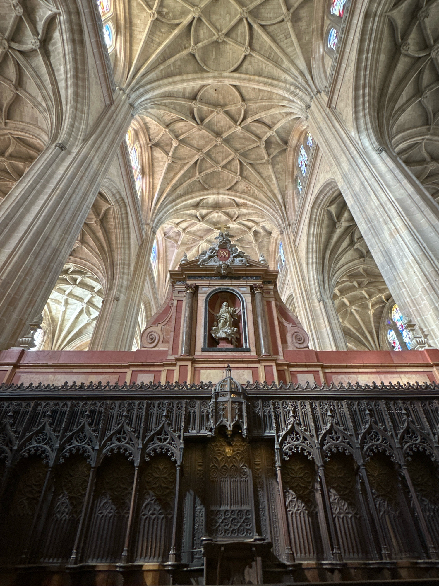The image depicts the interior of a grand Gothic cathedral, characterized by its soaring vaulted ceilings supported by massive stone pillars. The ceiling features intricate ribbed vaulting with complex geometric patterns, creating a sense of immense height and architectural complexity. Stained glass windows, visible in the upper portions of the image, cast colorful light onto the stonework. In the foreground, a section of ornate, dark wood choir stalls or a screen is prominently featured, displaying detailed Gothic tracery and architectural motifs. Behind the stalls, a decorative altar or reredos with a niche containing a sculpted figure is visible. The overall impression is one of historical grandeur and religious solemnity. This cathedral is likely the Cathedral of Segovia, located in the city of Segovia, which is approximately 30 kilometers from La Lastrilla, Spain. The image does not contain any people or specific actions.