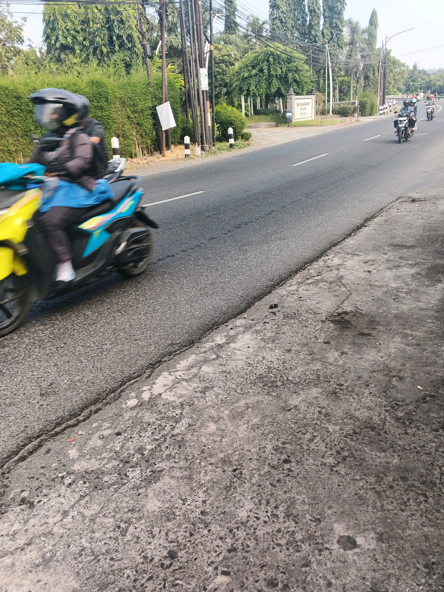 This image captures a scene on a road in Ungaran, Indonesia, likely during daylight. In the foreground, a person on a yellow and blue scooter is in motion, their figure blurred by the movement and possibly the camera's shutter speed. The rider wears a dark jacket and a black helmet with a clear visor. They are dressed in dark pants and a blue garment covering their legs. The scooter has a black seat and black wheels, with visible suspension.
The road itself appears to be paved asphalt with white lane markings. To the left of the road, a dense, green hedge forms a barrier. Beyond the hedge, tall trees line the background, their foliage creating a textured green canopy against a bright, possibly hazy sky. Several utility poles with numerous wires are scattered throughout the scene, suggesting an urban or semi-urban environment.
Further down the road, two more individuals on motorcycles are visible, also in motion. The one in front is wearing a blue shirt and a helmet, while the one behind wears a red shirt and a helmet. In the middle ground, a signpost stands on the left side of the road, and a white sign with text and decorative elements is mounted on a pole. The right side of the road shows a rough, textured concrete surface, indicative of a sidewalk or embankment. The overall impression is of a typical Indonesian road with active traffic and surrounding greenery.