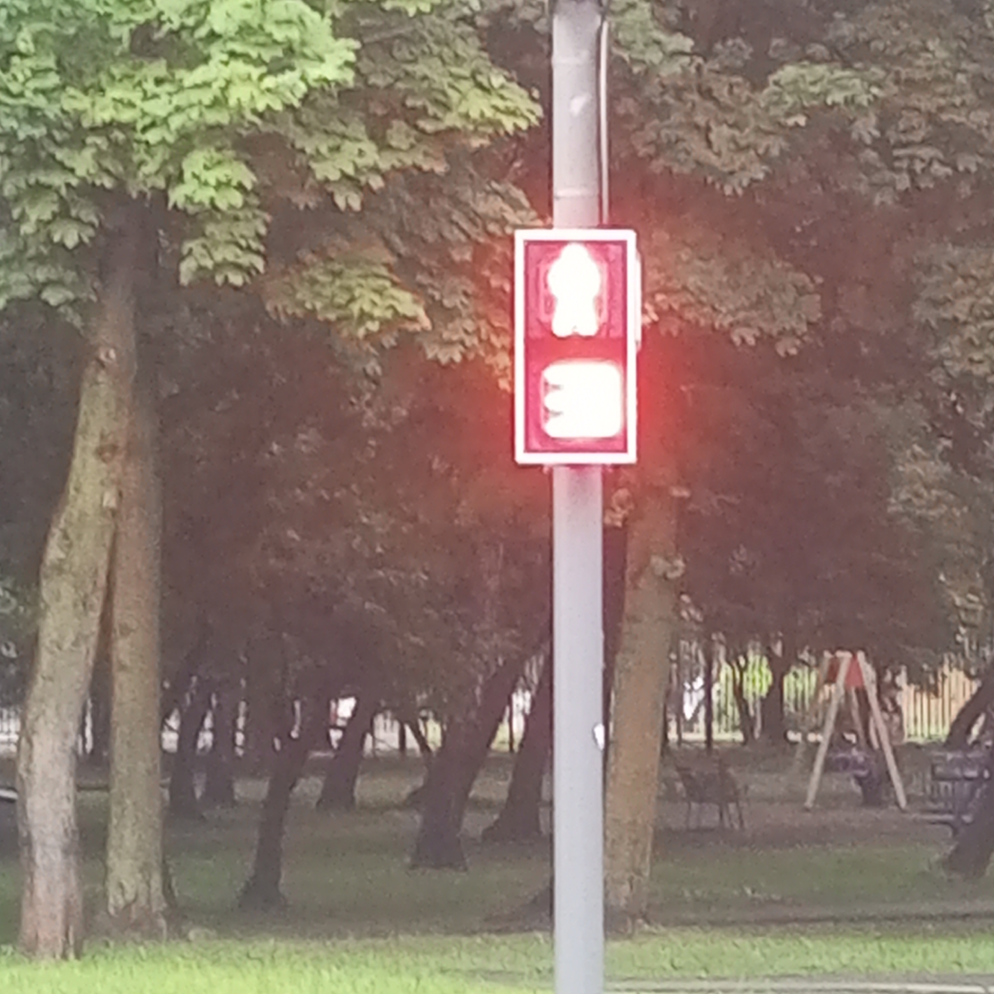 The image shows a traffic light with a pedestrian signal in a park setting. The pedestrian signal displays a white silhouette of a person on the top light and a white silhouette of a walking person on the bottom light, indicating "walk" for pedestrians. The traffic light is mounted on a grey pole and is surrounded by trees with green leaves and grass. In the background, there are more trees, some benches, and a playground with a red and white swing set. The overall scene suggests a public park, likely in a city. While the image does not provide specific location markers for Moscow, Russia, the presence of such public amenities is common in urban environments. There are no people visible in the immediate vicinity of the traffic light.