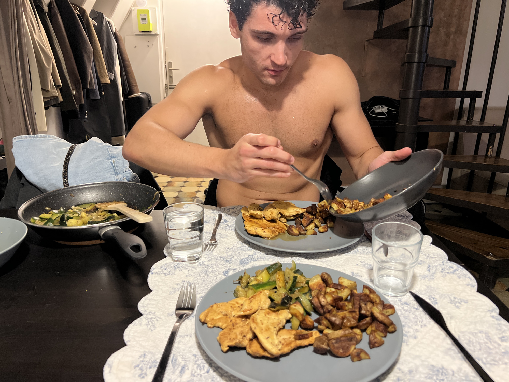In a home setting, a shirtless man with curly dark hair sits at a dark table, focused on transferring food from a pan to his plate with a fork. In front of him are two plates of food, each containing chicken strips, roasted potatoes, and sauteed zucchini. To the left, a frying pan with remaining zucchini sits on the table. A glass of water and cutlery are also visible. In the background, a coat rack with several jackets and a spiral staircase suggest a multi-story dwelling. The flooring is tiled. The lighting is warm and casts shadows, creating an intimate atmosphere. The scene does not provide specific details to indicate a location within Paris, France.