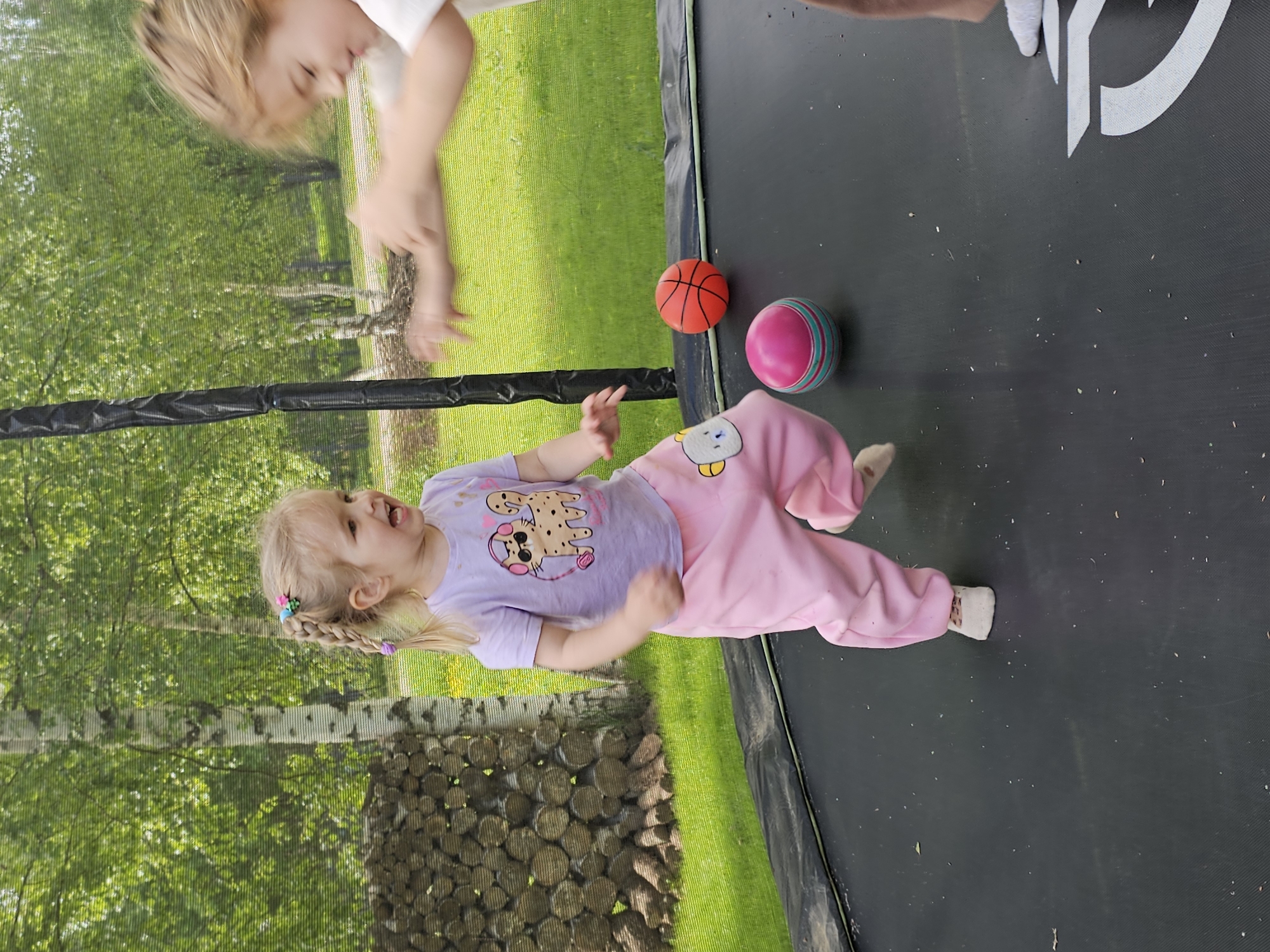 A young girl with blonde braided hair and a pink outfit is lying on her side on a black trampoline. A second, blurry child is partially visible near the top of the frame. An orange basketball and a pink-and-blue striped ball rest on the trampoline near the girl. The trampoline is surrounded by a safety net. Outside the trampoline, there is green grass and trees. A stack of firewood is visible in the background. There is no evidence this photograph was taken in the city of Мышкино, Russia.