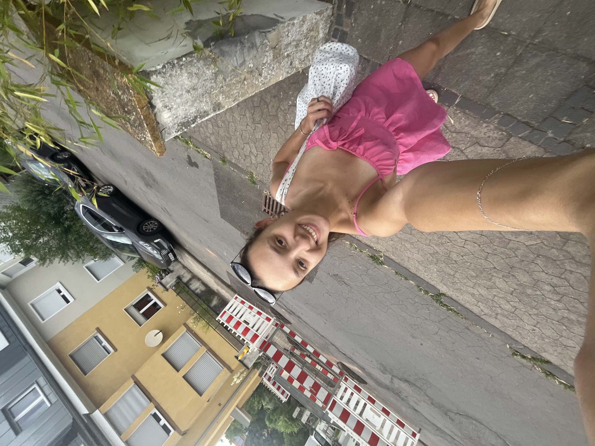 A selfie is taken by a woman with dark hair, sunglasses on her head, and a bright pink dress with spaghetti straps. She is smiling and holding a white patterned bag over her left shoulder. She is walking on a sidewalk next to a street where two dark-colored cars are parked. In the background, there are apartment buildings. Red and white construction barriers are visible on the side of the road. The scene appears to be in an urban environment. The architectural style of the buildings suggests it could be in Europe. The presence of construction barriers and the condition of the road indicate ongoing work or repairs. The overall lighting suggests it is daytime, possibly late afternoon or early evening.