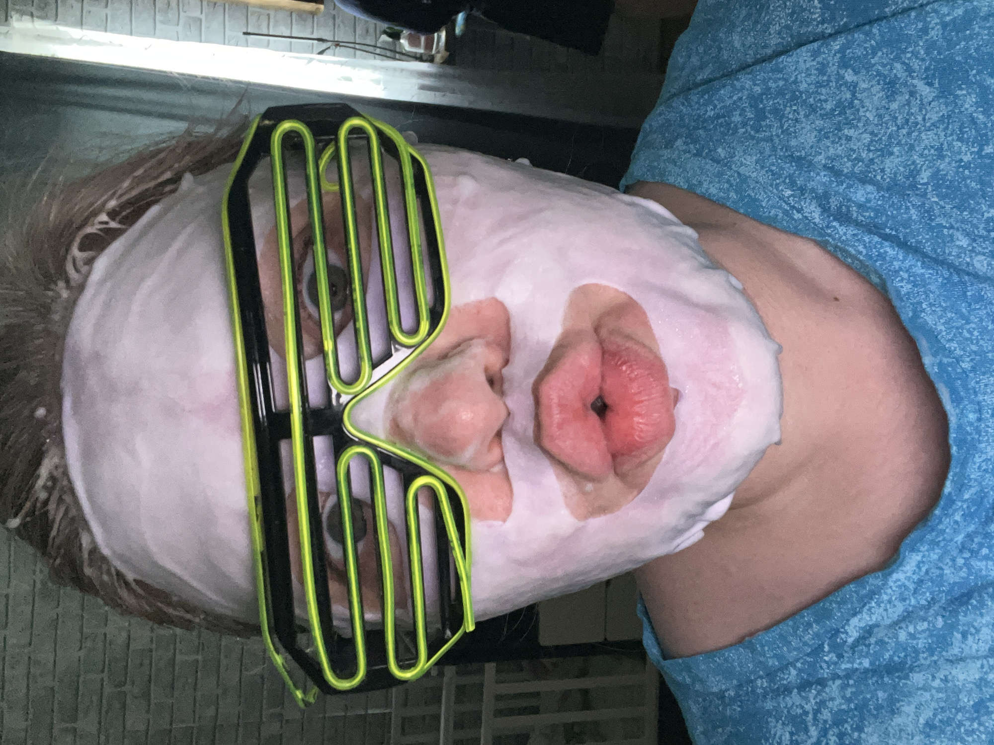 An individual is depicted lying down, with their head and upper torso angled towards the left side of the frame. Their face is entirely covered in a thick, white, foamy substance, likely a face mask or shaving cream, leaving only the eyes and mouth area partially discernible. Over their eyes, they wear distinct neon green and black slatted novelty glasses, often referred to as "shutter shades." The person's lips are prominently pursed, forming a "kissy face" or "duck face" expression, creating a humorous visual contrast with the foamy face covering. Light-colored hair is visible around the edges of the mask.
The individual is wearing a blue t-shirt with a subtle speckled pattern. The background suggests an indoor setting, possibly a bathroom or personal space, given the activity. In the upper left, a wall featuring a light-colored brick or tile pattern can be seen. A long, linear light fixture, resembling a fluorescent tube, extends across the top of the scene, indicating artificial lighting. Other parts of the background are darker and less distinct. The image is oriented with the person's head on the left and their body extending to the right, implying a 90-degree rotation. The overall impression is one of playful self-care or a lighthearted moment. There is no visible text in the image. This scene takes place in Vesyoloye, Russia.