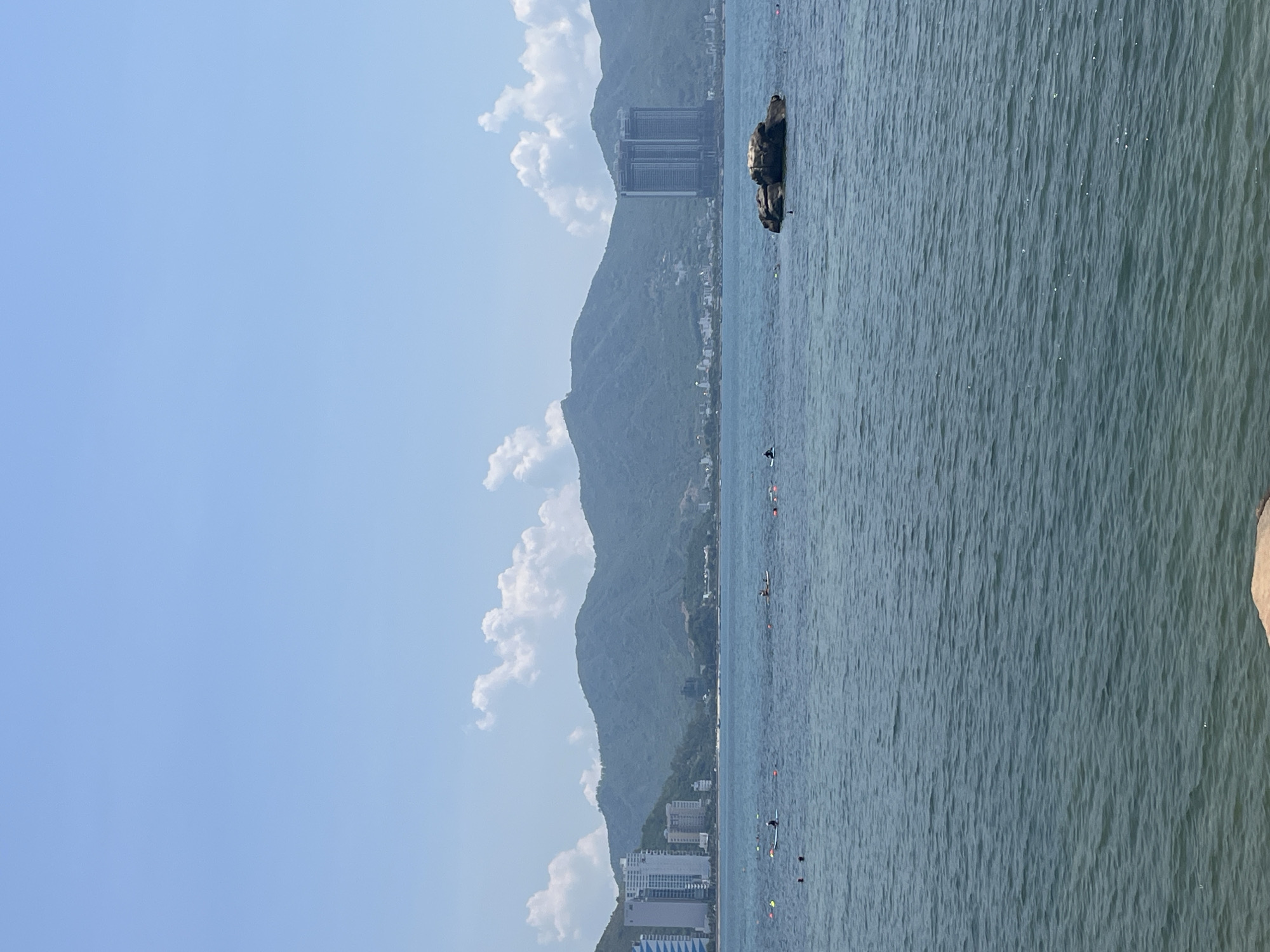 The image depicts a coastal scene in Bac Nha Trang Ward, Vietnam. The water in the foreground is calm with gentle ripples, reflecting the clear blue sky. Several buoys are visible in the water, indicating a swimming or recreational area. A few individuals are present in the water, some appearing to be on paddleboards and others swimming. In the mid-ground, a rocky outcrop emerges from the sea. The background features a long stretch of coastline with lush green mountains. Along the shore, modern high-rise buildings are interspersed with smaller structures, suggesting a developed urban area. The sky is bright and clear with scattered white clouds.