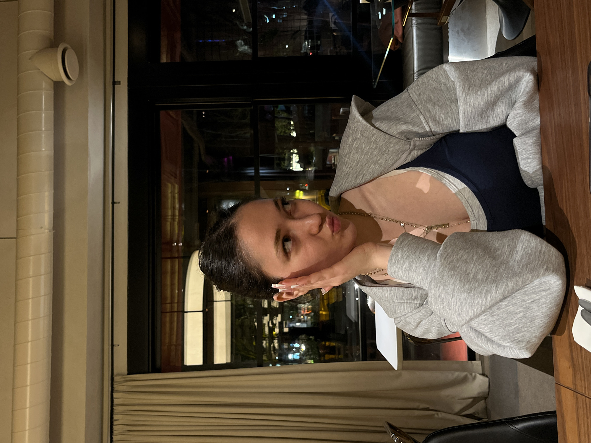 A woman is sitting at a table inside a restaurant, as seen from the reflection of city lights in the window behind her. She wears a dark-colored top with a light gray hoodie, resting her chin on her hand. The location is Yerevan, Armenia, based on the city lights reflected in the background. The interior features beige curtains, a wooden table, and a black chair. Pipes are visible along the wall to the left of the window.