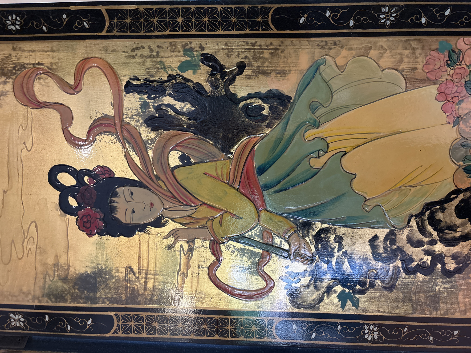 This is a detailed, close-up image of a decorative panel, likely a piece of artwork or screen, featuring a stylized depiction of an Asian woman. The artwork is rich in texture and color, with a prominent gold leaf background that shows some wear and patina. The central figure is a woman with dark hair styled elaborately with red floral accents. She wears a yellow robe with red trim around the waist and a green skirt. A flowing, translucent orange ribbon drapes around her and streams upwards. Her skin tone is light, and her facial features are delicately rendered. She appears to be holding a long, slender object, possibly a staff or brush, in her hands, with her fingers extended in a graceful pose. To the right of the woman, there is a dark, gnarled, tree-like form. The foreground at the bottom right shows a cluster of pink and coral flowers with green leaves. The panel is bordered on the left and right by dark, ornate vertical strips decorated with geometric patterns and floral motifs in gold and white. The overall impression is one of traditional Asian art, possibly from China or Japan, with a rich, aged aesthetic. The location in Chalong, Thailand is not discernible from the image itself.