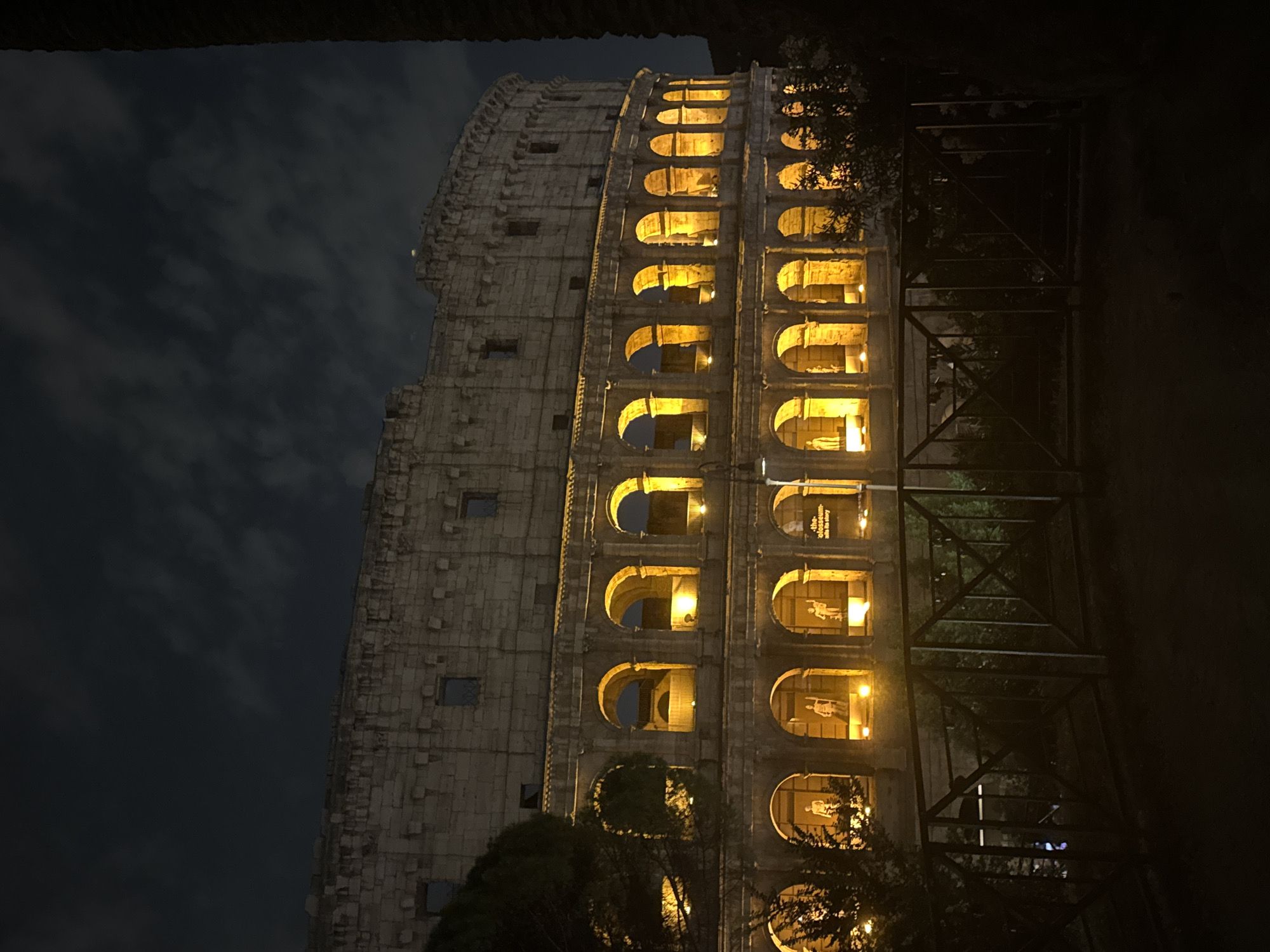 This nighttime photograph captures a vertical perspective of the illuminated Colosseum in Rome, Italy. The ancient amphitheater dominates the right and central portions of the frame, showcasing its iconic arcaded facade bathed in warm, yellow light. Multiple tiers of arches are visible, with many containing exhibition displays that feature silhouetted figures and informational panels. Faintly visible text within some of the illuminated arches reads "the Colosseum... its story," indicating an exhibition or interpretive display.
To the left of the brightly lit section, a darker, rougher part of the Colosseum's exterior wall rises against a dark, cloudy night sky. Scattered small, dark rectangular openings are visible on this less illuminated section. In the far right foreground, a dark, angular metal scaffolding or support structure partially obscures the view of the monument, blending with the shadows of nearby trees and foliage. The image conveys a majestic and historical ambiance, highlighting the architectural grandeur of the landmark under a dramatic night sky. No specific human activity or event is depicted, focusing solely on the monument itself.